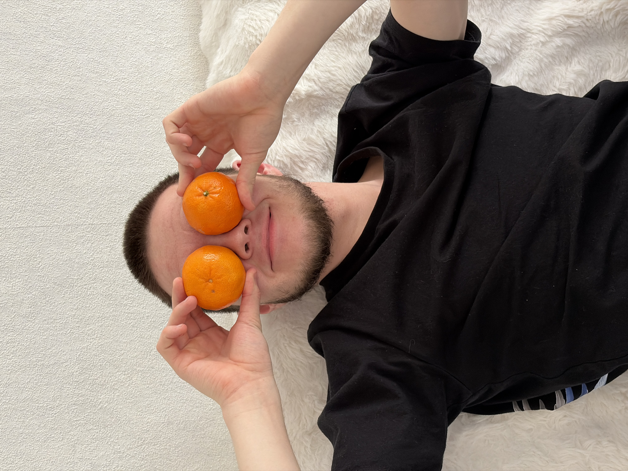 A man lies on a white, fluffy surface, possibly a rug or blanket. He is wearing a black long-sleeved shirt. He is holding two mandarin oranges up to his eyes. The location is Unknown, Unknown.