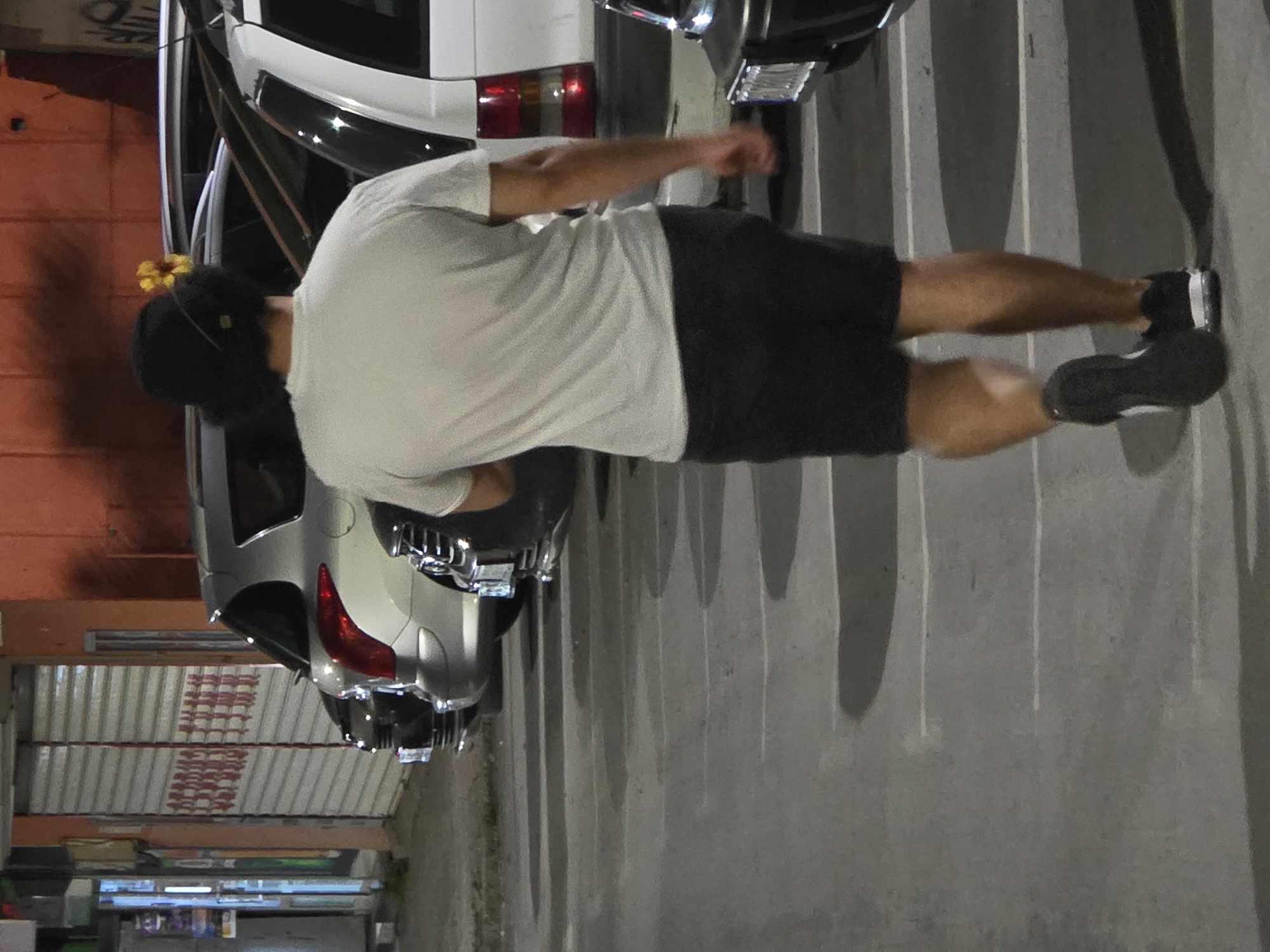 The image depicts a person walking away from the camera in a parking lot, possibly in Varna, Bulgaria. The individual is wearing a white t-shirt, dark shorts, and black sneakers. A yellow flower is visible tucked into the back of their dark, curly hair, which is covered by a dark cap. The person is walking on a paved surface marked with white parking lines. Several cars are parked in the background, including a silver SUV and a white van. The scene is illuminated by artificial light, suggesting it is nighttime. The background shows a building with a red-orange facade and closed metal roll-up doors, which may indicate a storefront or commercial establishment. The overall impression is a casual, everyday scene in an urban environment.