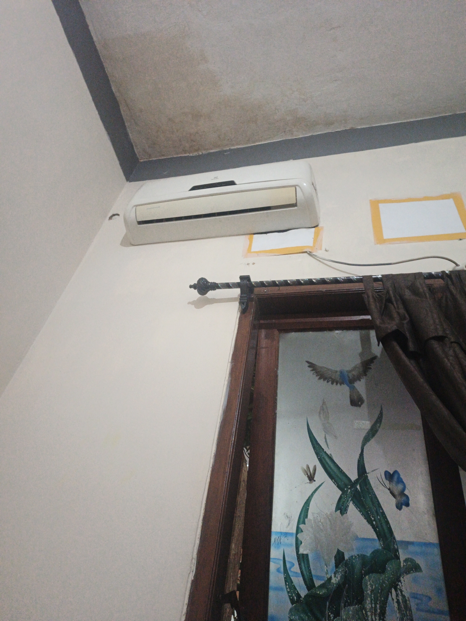 The image depicts an indoor scene, likely a room in a residential building, in Surabaya, Indonesia. A white air conditioning unit is mounted on a light-colored wall, near the ceiling. The ceiling itself appears to have some water stains or discoloration. Below the air conditioner, a dark brown curtain hangs from a curtain rod. To the right of the curtain rod, a wooden-framed window is visible, with decorative artwork featuring a bird in flight and aquatic plants and insects, including a blue butterfly. Yellow-taped squares are present on the wall beneath the air conditioner. The overall impression is that of a common living space.