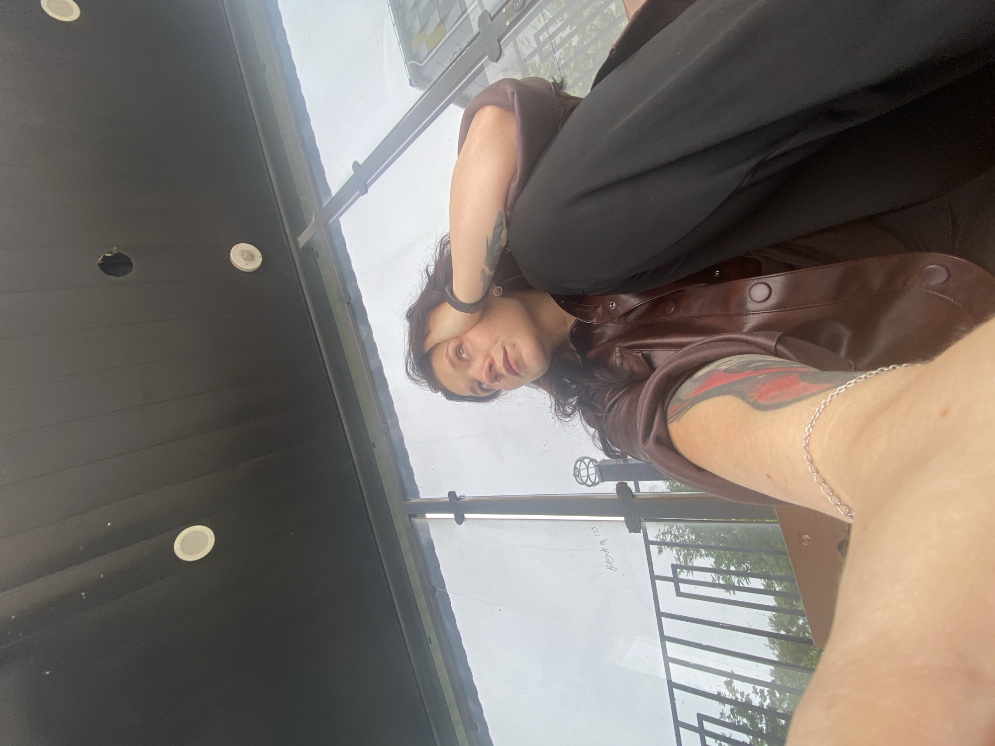 An indoor-outdoor selfie captures a young adult with dark, wavy hair, looking directly at the camera. The image is taken from a low angle, showing the person in a relaxed pose, with their body positioned diagonally across the frame. They are wearing a brown collared jacket or shirt, possibly leather, and black trousers. A black wristband is visible on their left wrist, and a thin silver chain bracelet adorns their right wrist. A prominent red and black tattoo is on their right forearm, while a darker tattoo is visible on their left forearm. One hand is gently placed near their face, partially obscuring their left eye. A nose piercing is also visible.
The setting appears to be a covered balcony or patio in Chaikovsky, Russia. Above, a dark grey or black paneled ceiling features two white circular recessed lights, with one light seemingly missing or having a hole next to it. A large glass barrier or window separates the person from the outside environment. Through the glass, an overcast sky is visible, along with blurred green trees and part of another building in the distance. A black metal railing with a decorative, spherical finial is also seen through the glass. Faint white text is visible on the glass barrier, reading "ВЯТЦКАЯ 133" (VYATSKAYA 133). The weather outside appears cloudy or overcast.