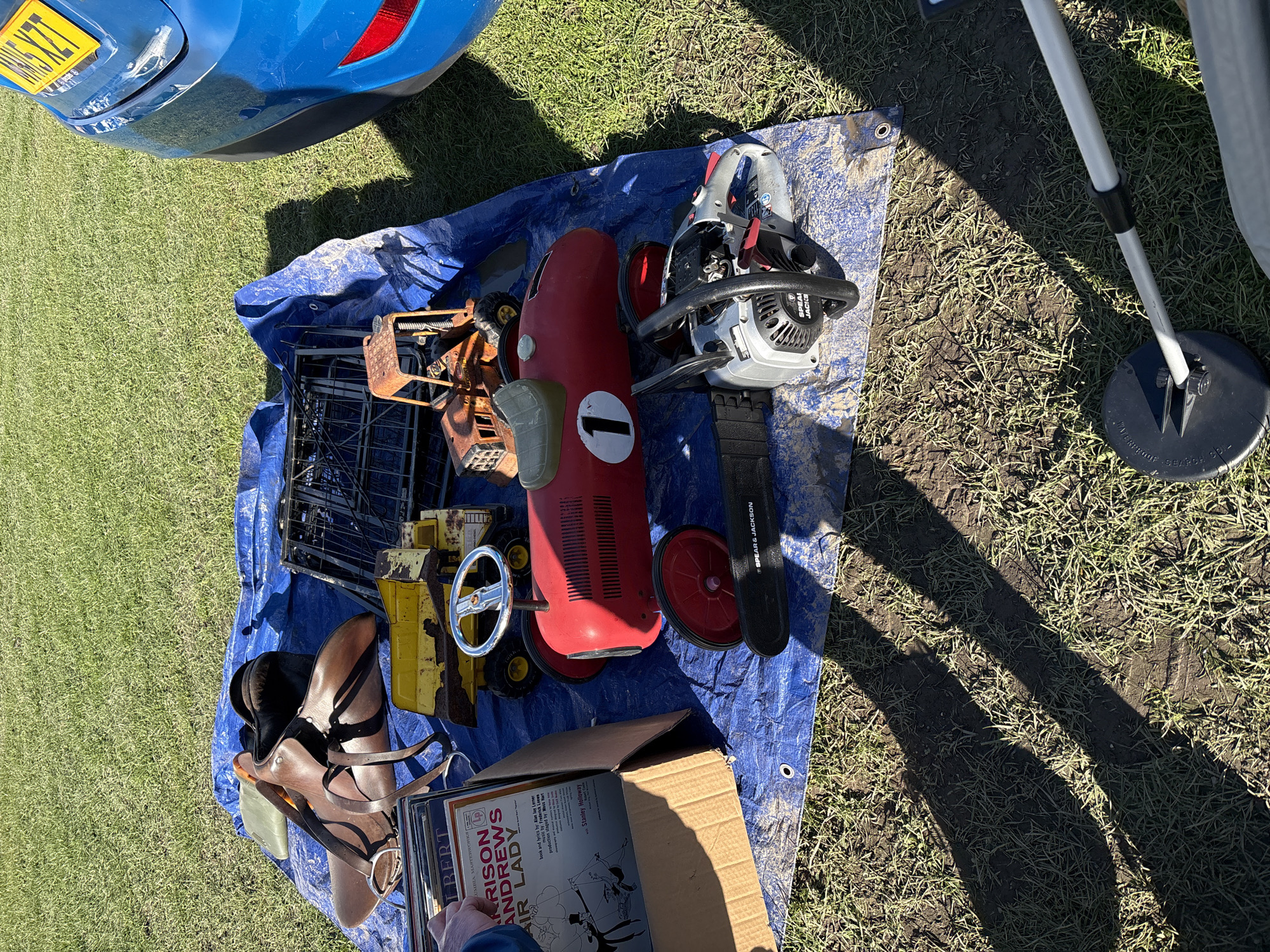 The scene depicts a collection of miscellaneous items spread out on a blue tarp laid on a grassy surface. A portion of a blue vehicle is visible in the top left. Prominent objects include a red pedal car with a "1" on its side, a chainsaw (Spear & Jackson), a metal cage, a yellow toy construction vehicle, a brown leather saddle, a box of vinyl records (including one titled "My Fair Lady"), and a metal detector. The tarp and items are placed on grass.
