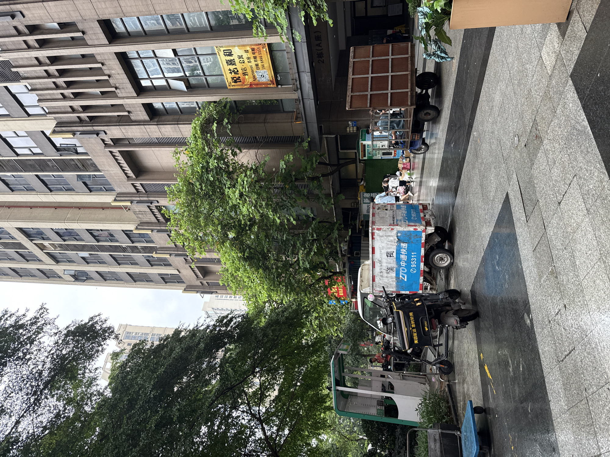 In Haizhu District, China, on a rainy day, a street scene unfolds with a backdrop of modern buildings. In the foreground, the wet pavement reflects the surrounding environment. Several vehicles are parked or in motion. A blue and white delivery truck with "ZTO EXPRESS" branding is prominently displayed, along with Chinese characters and a phone number "95311". Beside it, a black electric scooter with a yellow logo and Chinese text is parked. Further left, a small, green and white kiosk-like structure stands. In the middle ground, a three-wheeled truck with a wooden cargo bed is carrying people. A group of individuals, including adults and children, are visible near the kiosk and the delivery truck, some appearing to be engaged in activities or conversations. Trees with dense green foliage line the area, softening the urban landscape. In the background, a tall, multi-story building with numerous windows dominates the skyline. A yellow banner with Chinese text is affixed to the building's facade, advertising "Apartment Hotel".