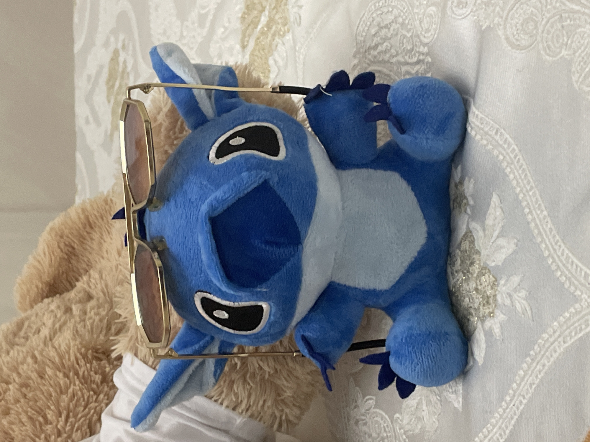 A blue plush toy resembling Disney's Stitch is prominently featured, lying on a white, intricately textured surface, likely a bed or pillow, in an indoor setting. The Stitch plush toy is adorned with a pair of gold-framed sunglasses with brown-tinted lenses, resting on its head and ears. Its characteristic large black and white eyes, open mouth, and blue fur with lighter blue patches on its belly and inside its ears are clearly visible. Small, dark blue claws are also evident on its paws. Part of a larger, light brown, furry plush toy is visible behind Stitch on the left side of the frame. The white fabric beneath the plush toys displays a decorative pattern, possibly embroidery or a textured weave, adding a refined touch to the background. The lighting suggests an interior environment, possibly during the daytime. No specific activity is taking place; the image captures a still arrangement of toys. No text is visible in the image. The scene is located in Ihtiman, Bulgaria.