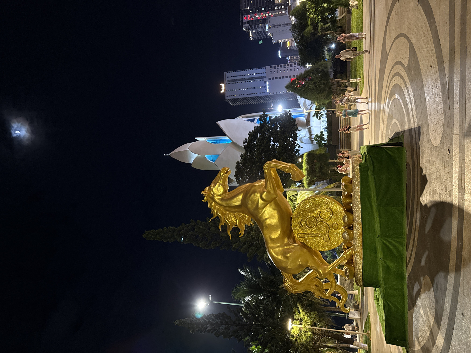 This is a low-angle, night-time photograph of a golden horse statue in the city of Nha Trang, Vietnam. The horse, depicted rearing on its hind legs, is positioned on a platform covered in green artificial grass. Beneath the horse, a large, ornate golden coin is visible, adorned with decorative elements and what appears to be a stylized symbol. Surrounding the base of the statue are small, golden ingot-shaped objects.
In the background, a cluster of modern buildings, including a tall, slender skyscraper with the "NOVOTEL" logo, dominate the skyline. A unique, lotus-shaped architectural structure with blue accents is also prominent. Trees, including tall cypress-like evergreens and palm trees, are scattered throughout the park-like setting. Streetlights illuminate the scene, casting shadows on the patterned pavement.
Several people are visible in the mid-ground, walking and standing in small groups. They are casually dressed, and some appear to be observing the statue. The sky above is dark, with a bright moon partially obscured by clouds. The overall impression is a festive and illuminated public space. This is the Horse and the Dragon statue, a popular landmark in Nha Trang, Vietnam.
