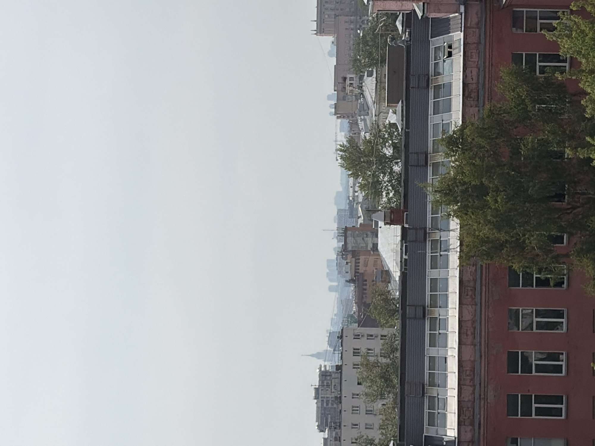 The image depicts a cityscape with several buildings and a hazy sky. In the foreground, there is a red-brick building with large windows. Above it, there is a darker-colored roof with a series of skylights. Behind this building, other structures of varying heights and architectural styles are visible, with some appearing to be older apartment buildings and others more modern constructions. In the distance, a tall, spire-like building stands out, characteristic of Stalinist architecture, likely the Ministry of Foreign Affairs building on Smolenskaya Square in Moscow, Russia. The overall atmosphere suggests a slightly overcast or smoggy day, typical of urban environments. Trees are interspersed between the buildings, adding a touch of nature to the concrete landscape.