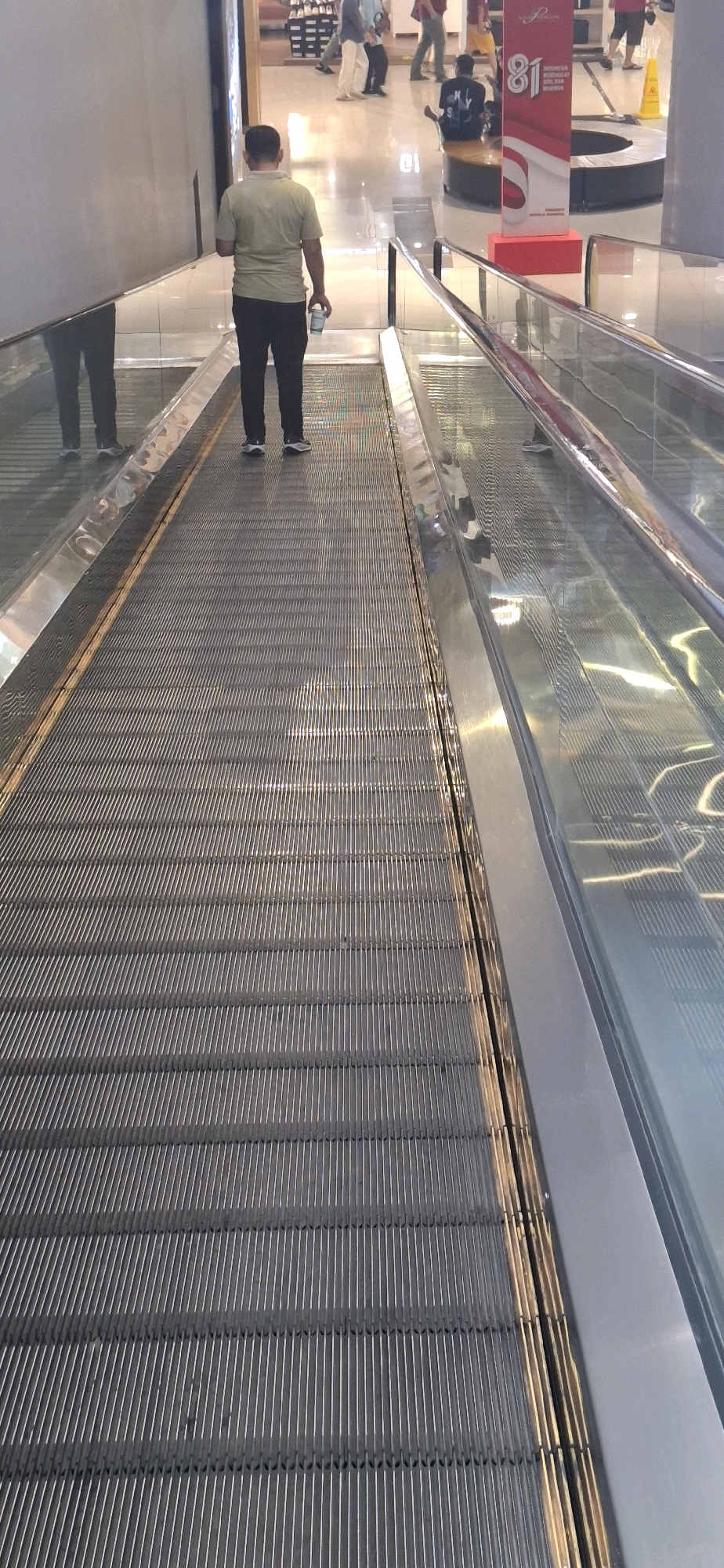 In Surakarta, Indonesia, a man is captured from behind on an escalator, descending. He is wearing a light green polo shirt and dark trousers, holding a white cup in his right hand. The escalator's metal steps are clearly visible, with a reflective glass barrier to the right. In the background, other individuals are present, with a red and white promotional banner for "81 Indonesia Merdeka" visible on the right side of the frame. The environment appears to be an indoor shopping mall.