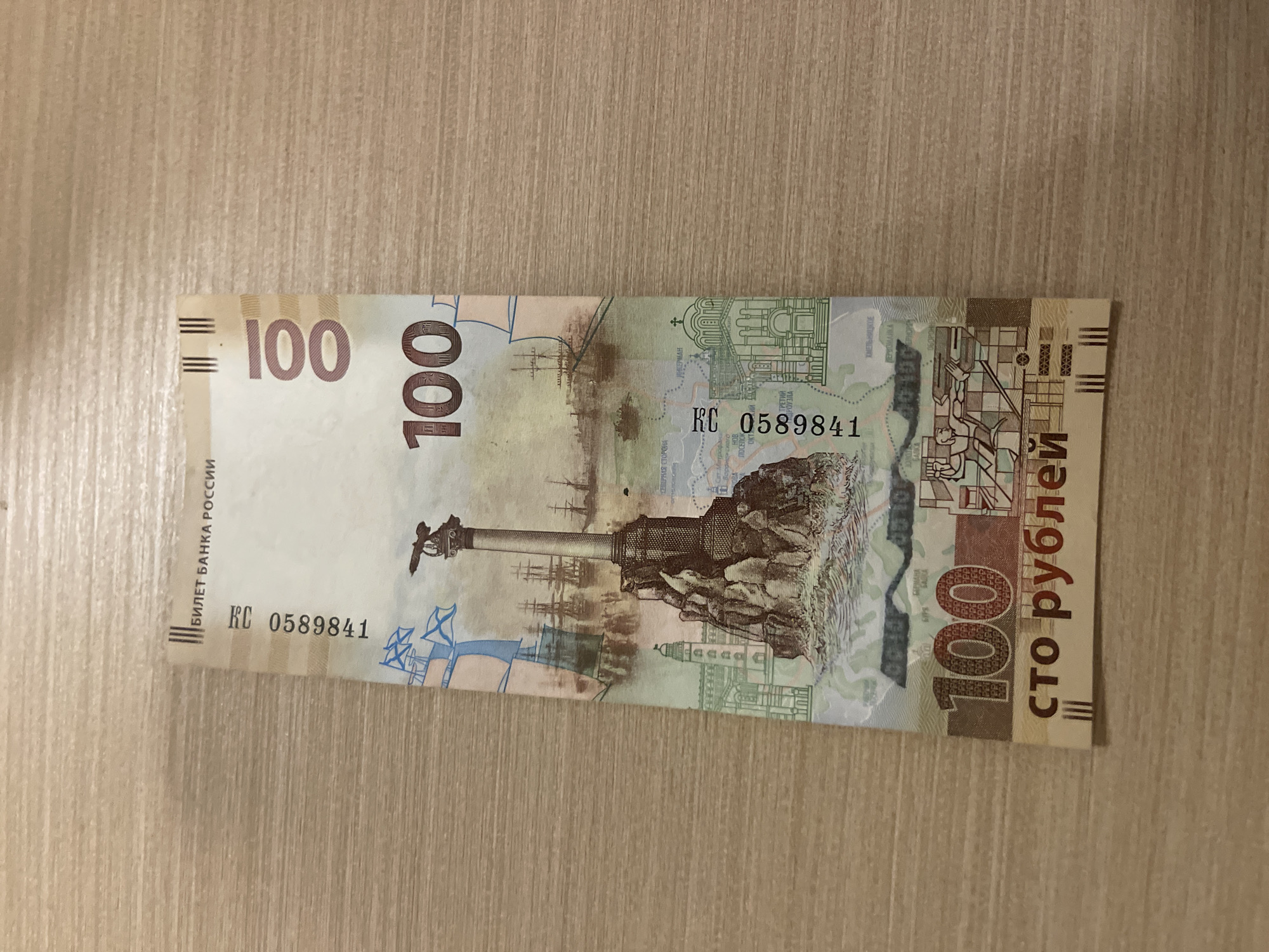 The image shows a Russian 100 ruble banknote lying on a wooden surface. The banknote features a depiction of the Monument to the Sunken Ships in Sevastopol, a lighthouse, and sailing ships. The text "БИЛЕТ БАНКА РОССИИ" (Ticket of the Bank of Russia) is visible at the top, and "100" is printed multiple times. The serial number "КЦ 0589841" is also present. The bottom of the banknote shows the text "сто рублей" (one hundred rubles). There is no indication of Panyu District, China, in the image.