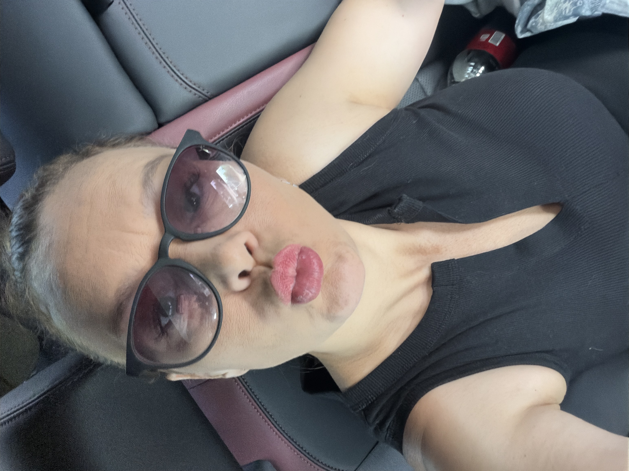 A woman is taking a selfie in a car, leaning slightly to her right. She is wearing a black tank top and dark sunglasses. Her lips are pursed, and she is looking directly at the camera. The interior of the car is visible behind her, with dark gray and red seating. There is a red object, possibly a bottle or can, in the background to her right, next to a crumpled gray fabric. The lighting suggests it is daytime. There is no indication of the location within the city of Chaikovsky, Russia.