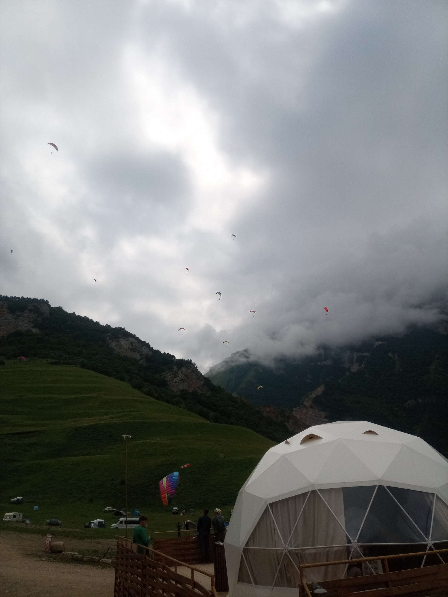 This image depicts a paragliding event at El'tiubiu, Russia, under a cloudy sky. In the foreground, a white geodesic dome structure with triangular window panels is visible, along with a wooden railing and a few people standing near it. The middle ground features a green, grassy slope with several vehicles, tents, and a colorful paraglider suspended mid-air. The background is dominated by a steep, tree-covered mountain range partially obscured by clouds. Numerous paragliders are scattered across the sky, indicating an active paragliding session. The overall atmosphere appears to be a recreational outdoor activity in a mountainous region.