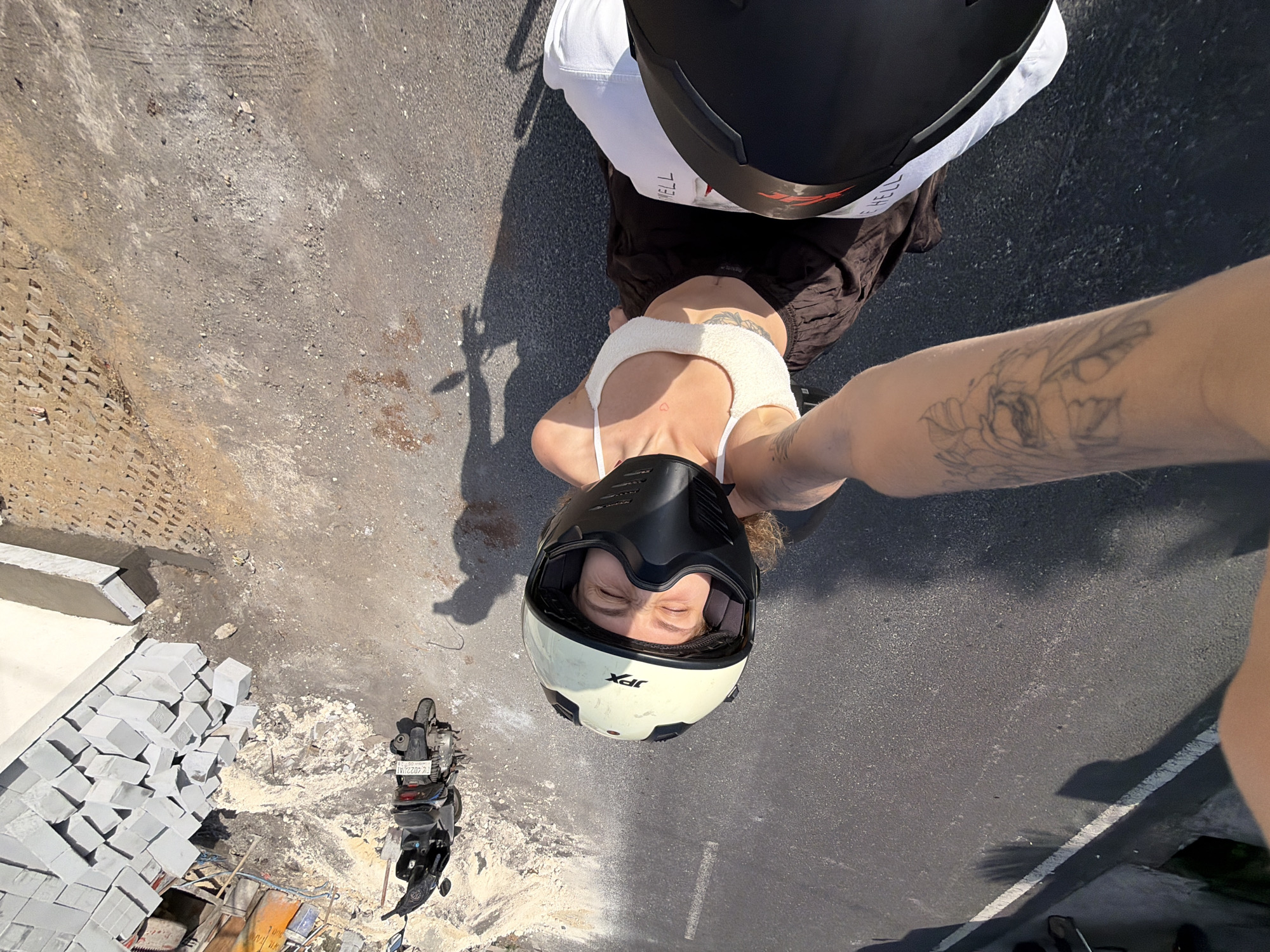 This is a first-person perspective, high-angle shot of two individuals on a motorcycle. The scene appears to be a construction site or an area undergoing development, with stacks of gray rectangular blocks visible on the left. The ground is a mix of asphalt and dirt.
The primary subject is a person wearing a white crop top, dark brown shorts, and a white and black helmet. Their eyes are closed, suggesting relaxation or enjoyment. They have tattoos on their arm. Partially visible behind them is another person also wearing a helmet. A black motorcycle is parked nearby.
The time of day is daytime, indicated by the bright sunlight casting strong shadows. The weather appears to be clear and sunny.
Visible text includes "JPX" on the helmet and what appears to be "Fk 4022UJ" on the motorcycle's license plate. The setting, with construction materials and a motorcycle, implies an outdoor, possibly urban or suburban, environment in North Kuta, Indonesia. The overall mood seems casual and adventurous.