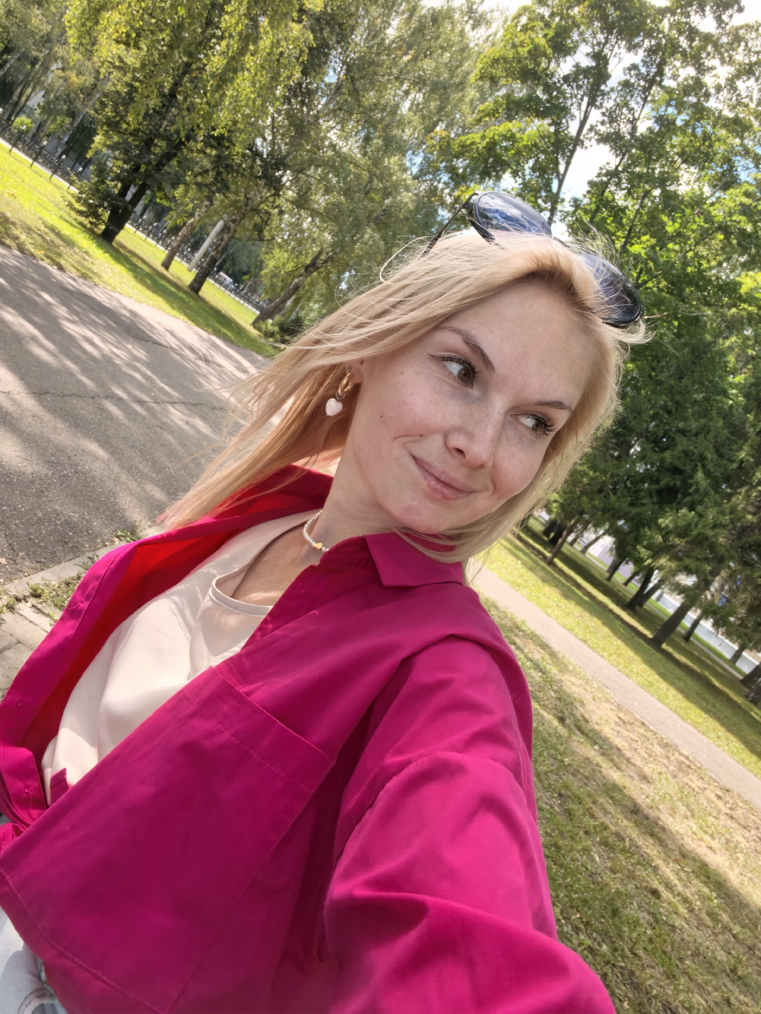 In Sterlitamak, Russia, a young blonde woman with freckles and light brown eyes smiles as she takes a selfie outdoors. She is wearing a bright pink shirt over a white top, a pearl necklace with a gold heart pendant, and heart-shaped earrings. Sunglasses rest on top of her head. She is positioned on a paved path, with a grassy area and trees in the background. The sun is shining, casting shadows on the path.