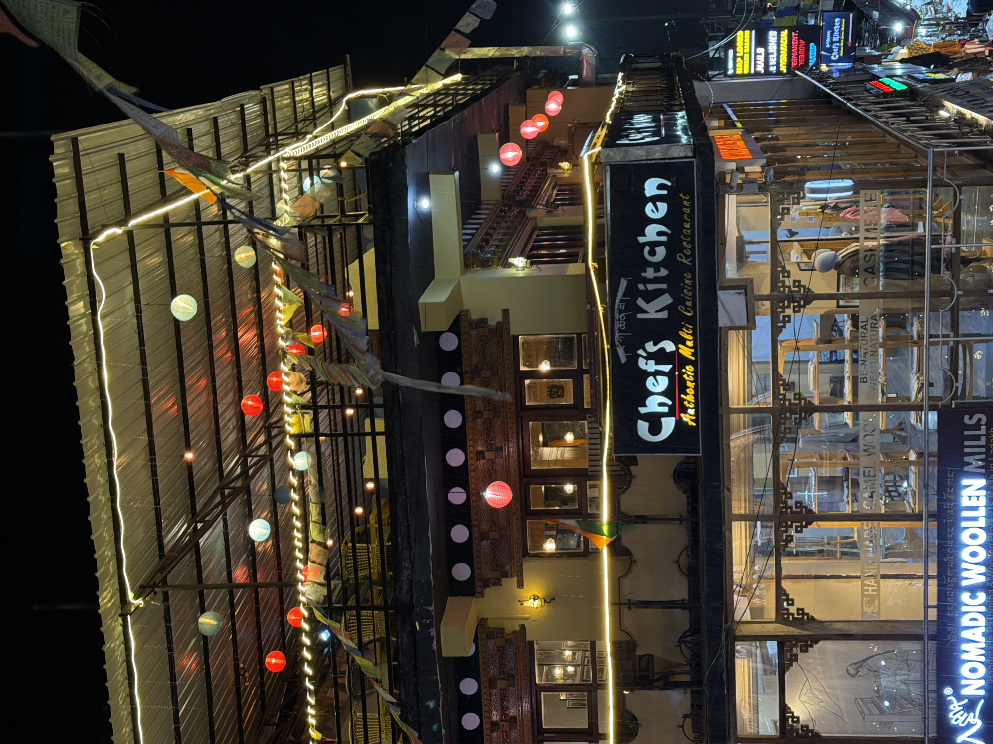 The image shows a multi-story building at night, likely in the city of Leh, India, given the Tibetan prayer flags and the sign for "Chef's Kitchen" with text that appears to be in Tibetan script. The building has a traditional architectural style with ornate wooden carvings around the windows. The upper floors are illuminated with string lights and red lanterns, creating a festive atmosphere. A prominent sign reads "Chef's Kitchen Authentic Multi Cuisine Restaurant." Below this, a shop window displays what appears to be woolen products, with a sign that reads "NOMADIC WOOLLEN MILLS." The street level shows other businesses with illuminated signs, suggesting a commercial area. The overall scene depicts a vibrant and well-lit commercial street at night.