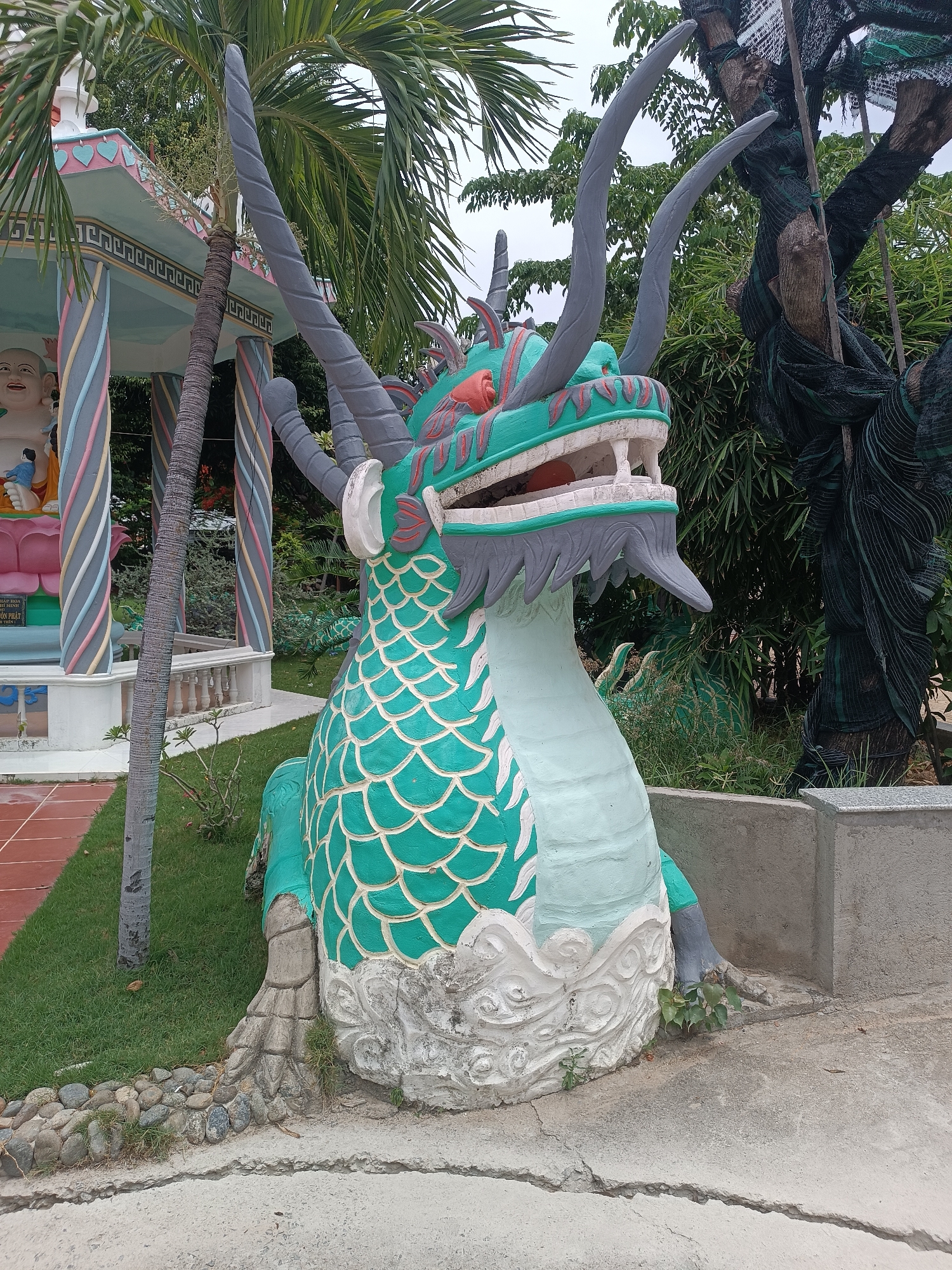 In Cam Ranh, Vietnam, an outdoor scene features a prominent, vibrantly colored statue of a dragon. The dragon statue, primarily painted in green and white with gray accents, is positioned in the foreground, facing slightly to the right. It has a detailed scaly body and a fierce expression with its mouth open, revealing teeth and a red interior. Its large, pointed horns curve upwards.
Behind the dragon, to the left, is a colorful pavilion with striped pillars in shades of pink, blue, and yellow. A statue of a cheerful Buddha figure, seated and smiling, is visible inside this pavilion. A palm tree with broad fronds stands between the dragon and the pavilion.
The ground around the dragon is a mixture of concrete, grass, and decorative stones. To the right of the dragon, there is a concrete planter or ledge, and behind that, dense greenery, including bamboo, and a tree covered in dark netting. The sky is overcast.