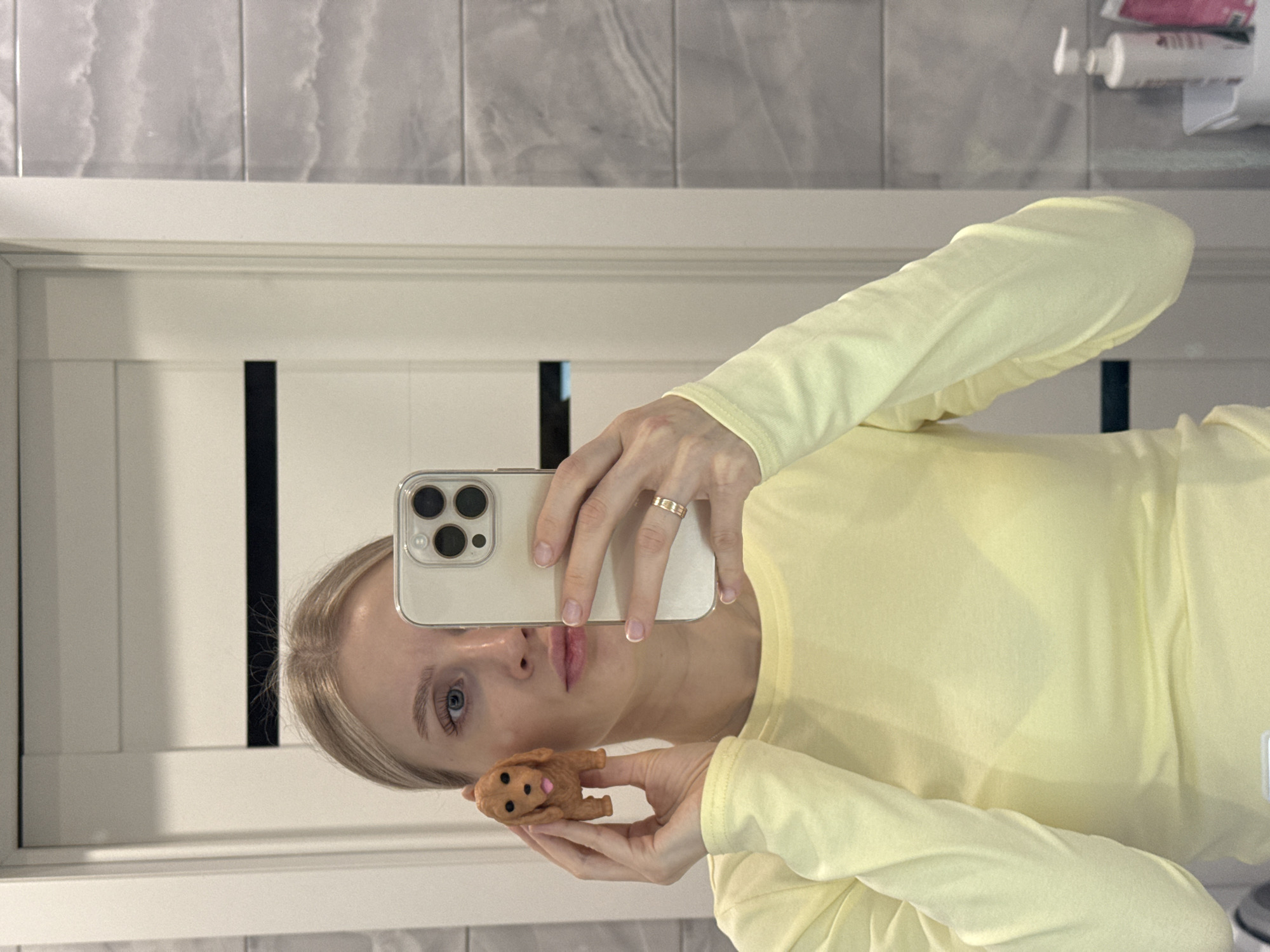 A person with blonde hair and blue eyes is holding a smartphone in their right hand and a small, brown, toy dog in their left hand. The smartphone is reflecting the person's face and upper body. The person is wearing a light yellow long-sleeved shirt. The background appears to be a bathroom with light gray tiled walls and a white door with black accents. Several bottles of what appears to be lotion or hairspray are on a shelf in the upper right corner of the frame. It's not possible to determine if the location is Izhevsk, Russia from the image alone.