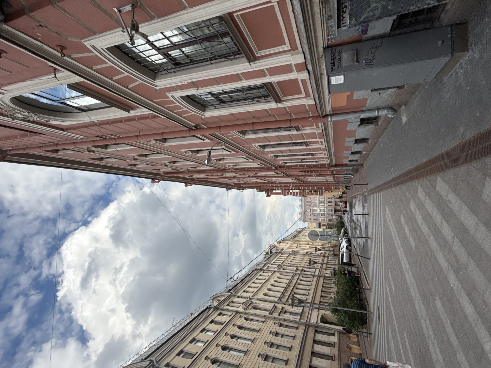 A portrait-oriented image captures an upward, slightly angled view of a city street flanked by multi-story buildings under a partly cloudy sky.
On the left, a prominent reddish-brown building features a classical facade with numerous windows framed by white decorative cornices and sills. Some windows are protected by security bars, and a network of pipes runs along the exterior. Reflections in the windows show portions of the blue sky and clouds.
On the right, a lighter, yellowish-cream colored building extends into the background, showcasing ornate architectural details and multiple windows. Attached to this building are dark grey utility boxes and pipes. Visible graffiti on one of the boxes includes fragmented letters such as "3MAPOBA" and "EMEST".
The street level is visible in the lower part of the frame, with a paved surface and distinct light-colored crosswalk stripes. Further down the street, a white car is parked, and a few blurred figures of people are discernible walking on the sidewalk. Green foliage from bushes or trees adds a touch of nature to the urban setting. A round traffic sign is also visible on the right.
The sky, occupying the upper-left portion, exhibits a mix of clear blue patches and large, billowy white cumulus clouds, indicating daytime. Utility wires or power lines crisscross the sky between the buildings. The scene depicts an active urban environment, possibly in an older European city, during daylight hours with fair weather.