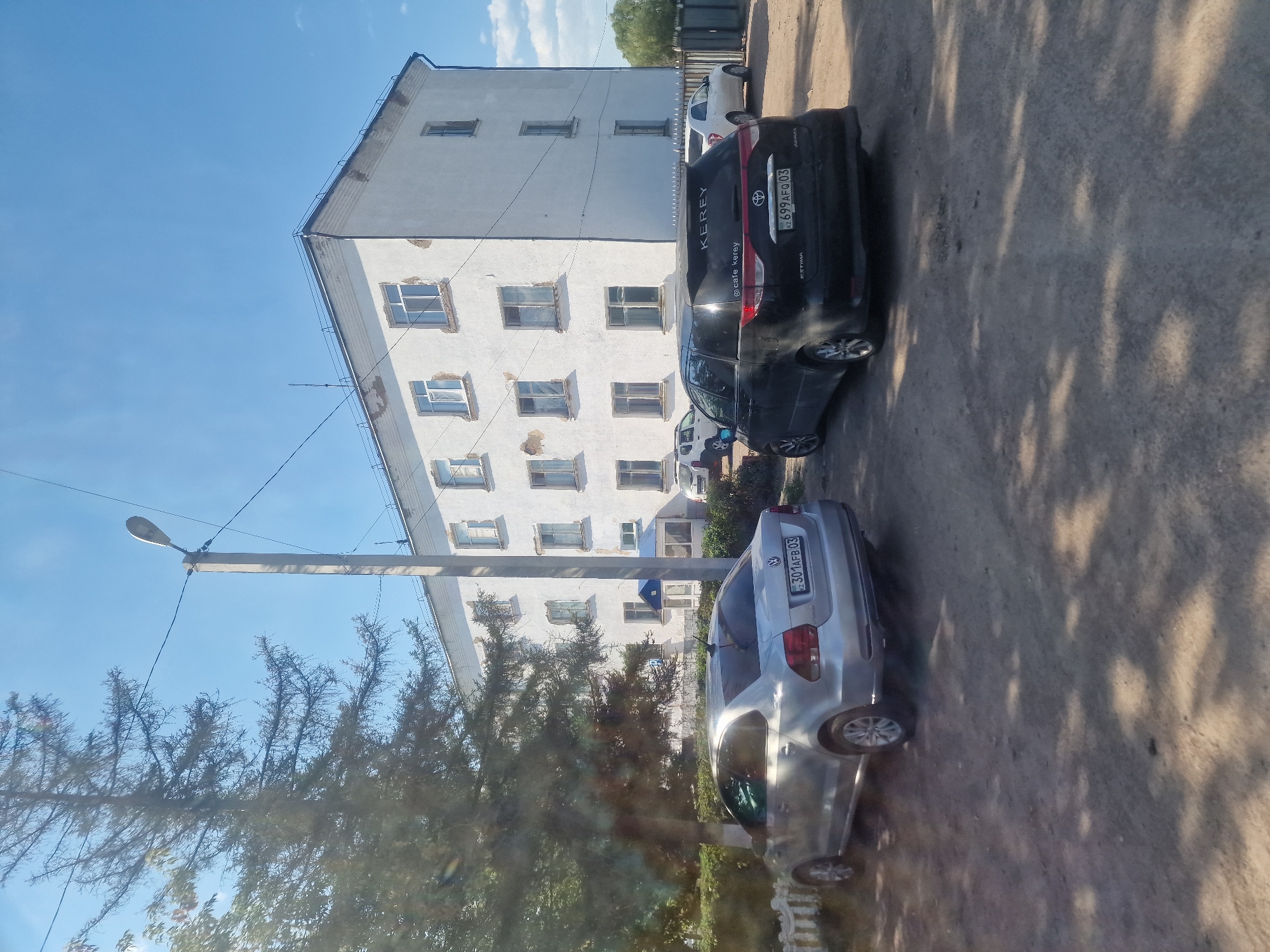 In the city of Makinsk, Kazakhstan, an image captures a sunny day with a clear blue sky. In the foreground, a silver Volkswagen sedan is parked on a gravel surface. To the right of the sedan, a black minivan is also parked. Behind the vehicles, a multi-story white building stands, its facade showing signs of wear. A tall, slender street lamp is visible on the right side of the image. Trees, including a large pine tree on the left, frame the scene. The image appears to be taken from a low-angle perspective, possibly from inside another vehicle, as evidenced by some reflections on the left side.