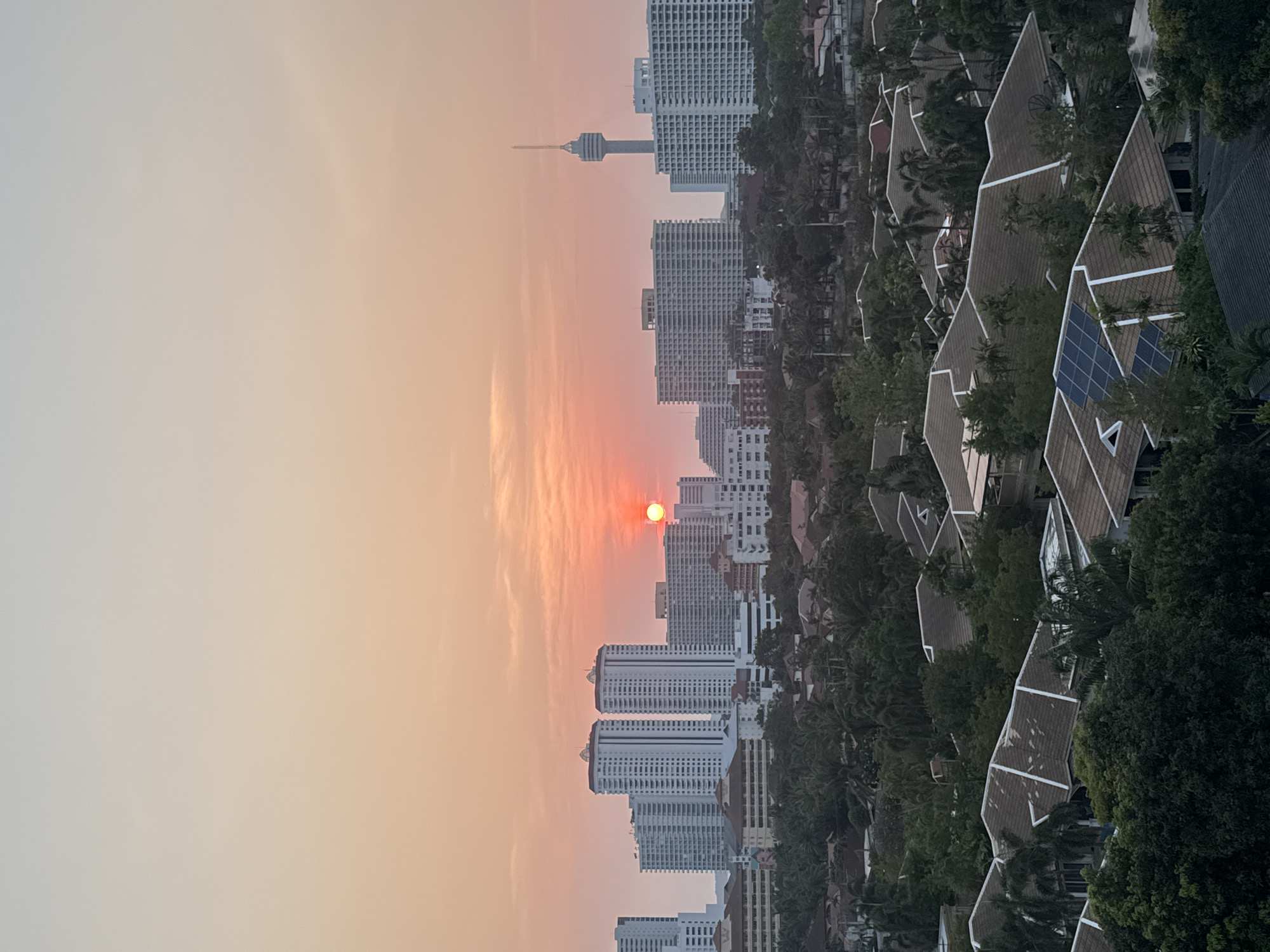 The image shows an urban landscape with a hazy, orange sky suggesting sunrise or sunset. A large, bright sun disc is visible through the haze, positioned over a cluster of high-rise buildings. These buildings are mostly white with multiple floors and a repetitive window pattern. A tall communication tower is located further in the background. To the right of the buildings, there are houses with steeply pitched roofs, partially obscured by trees. Some of the roofs appear to have solar panels. The city is Pattaya City, Thailand.