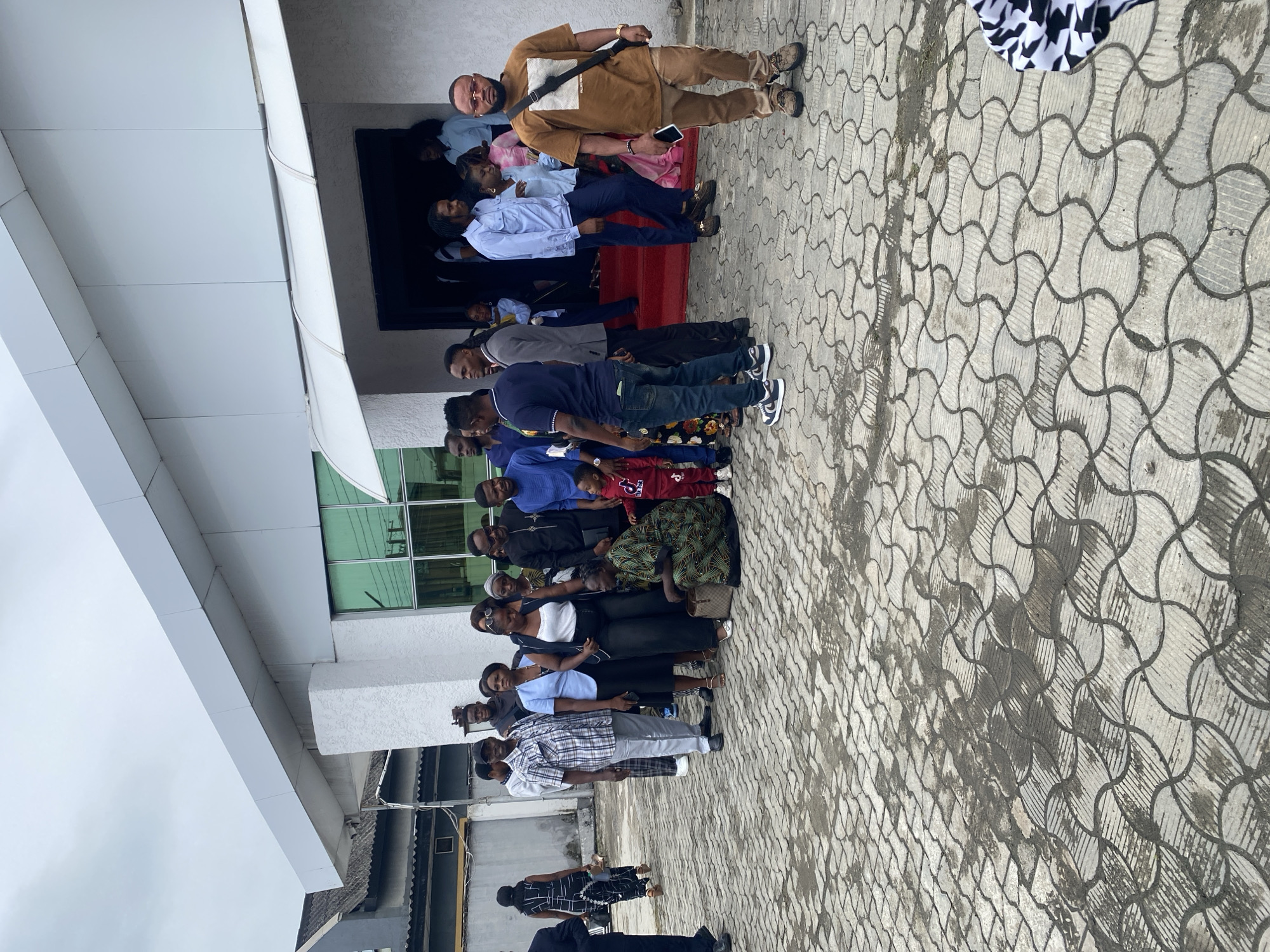 The image shows a diverse group of people standing in a line in what appears to be the city of Lekki, Nigeria. The background includes a building with a modern architectural style, featuring a white exterior, a flat roof, and green-tinted windows. The ground is paved with interlocking concrete blocks with a distinctive pattern. The group of people consists of both men and women of varying ages, dressed in a mix of casual and formal attire. One man in a brown outfit stands at the top of the steps. The people are standing closely together and appear to be waiting. The atmosphere appears calm and organized.
