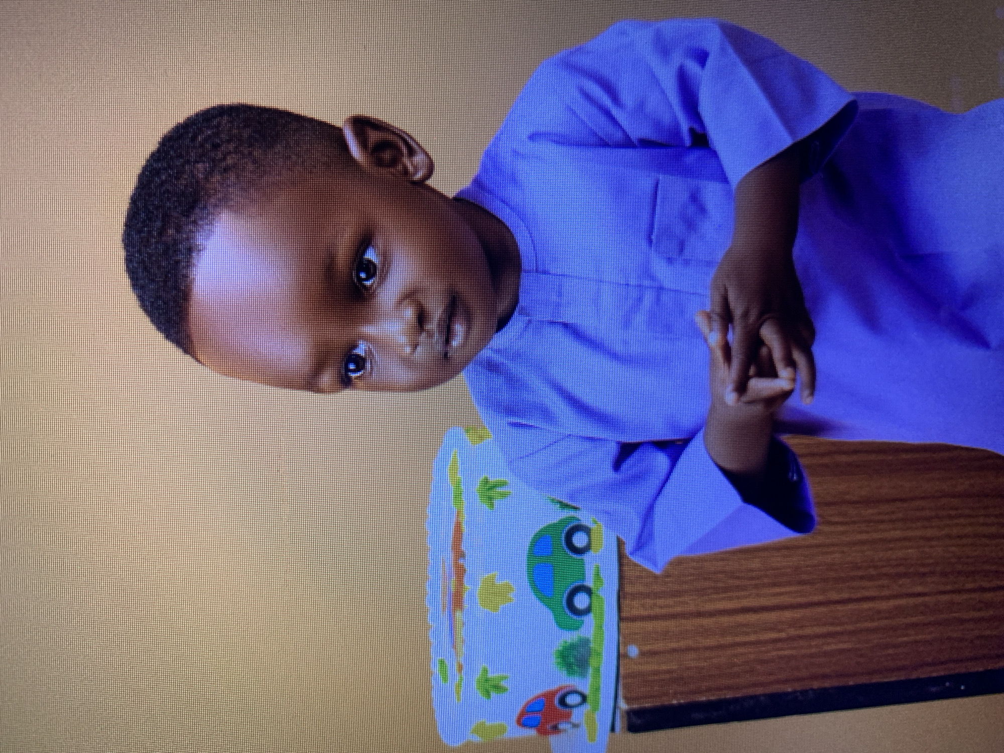 A young boy stands to the right of a cake decorated with cartoon cars. He is wearing a light purple, traditional African garment with long sleeves and a high neckline. His hands are clasped in front of him. The background is a solid, light brown color, and to the left of the boy is a wooden cabinet or stand, on top of which sits the cake. The cake is rectangular and white with various colorful cartoon cars, trees, and what appear to be clouds or abstract shapes. The overall scene suggests a celebratory event, possibly a birthday. The image quality indicates it was taken of a screen, likely a monitor displaying a photograph. The provided location of "Gura Top, Nigeria" is not visually ascertainable from the image itself.