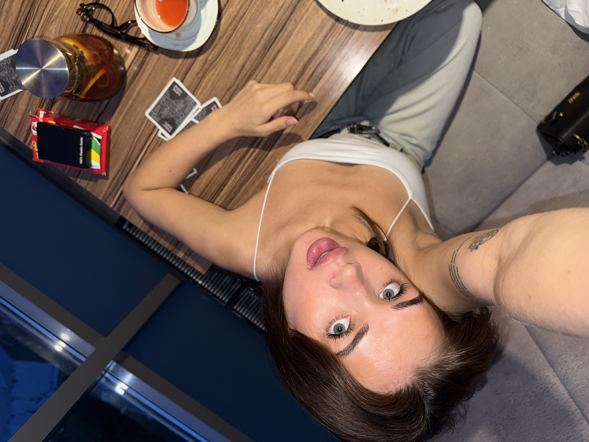 The image captures a female individual in an indoor setting, identified as being in the city of Perm, Russia. The perspective is from directly above or slightly below eye level, looking down at the subject who is reclining or sitting low on a grey upholstered sofa or bench.
The central figure is a young adult female with dark brown hair, light blue-green eyes, and full lips slightly parted, revealing a glimpse of her tongue. Her eyes are wide, and she is looking directly into the camera. She wears a white spaghetti-strap tank top and light-colored (possibly olive green or khaki) trousers. A small tattoo with text is visible on the inner bicep/forearm of her right arm, which is resting on the grey upholstery beside her head. Her left arm is partially visible, resting on a wooden table.
The wooden table, featuring a visible grain pattern, holds several items. These include a red box of "UNO 100% Plastic Cards" with a black card case on top, and several scattered playing cards with black backs marked "GAME STORE." A clear glass carafe with a metal lid contains a reddish-orange beverage with visible citrus slices and possibly mint leaves. Next to the carafe is a pair of black-rimmed eyeglasses. A white ceramic teacup with a saucer, containing a small amount of reddish-orange liquid, is positioned to the right of the carafe. A white plate with some food residue is partially visible on the far right edge of the table.
The background on the left features a large window with dark frames, reflecting blurred lights, suggesting a nighttime or dark exterior view. A dark blue wall or panel is adjacent to the window. Below the window, a dark metal radiator or vent is partially visible. The grey upholstered seating extends throughout the foreground and midground, with a small black handbag with a gold chain strap resting on it in the lower right corner. The lighting in the scene appears to be artificial and warm.