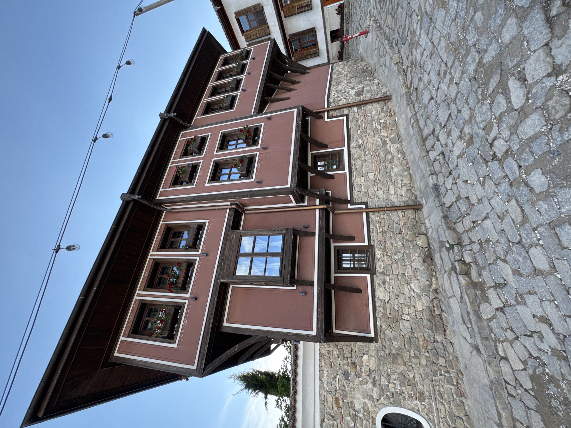 The image depicts a multi-story traditional Bulgarian house in Plovdiv, Bulgaria. The house features a reddish-brown exterior with white trim and dark wood accents. The windows are arranged in rows and have flower boxes with red flowers. The house has an overhanging upper level supported by wooden beams. The street is paved with cobblestones. Above the house, there are overhead light fixtures. The sky is blue.
