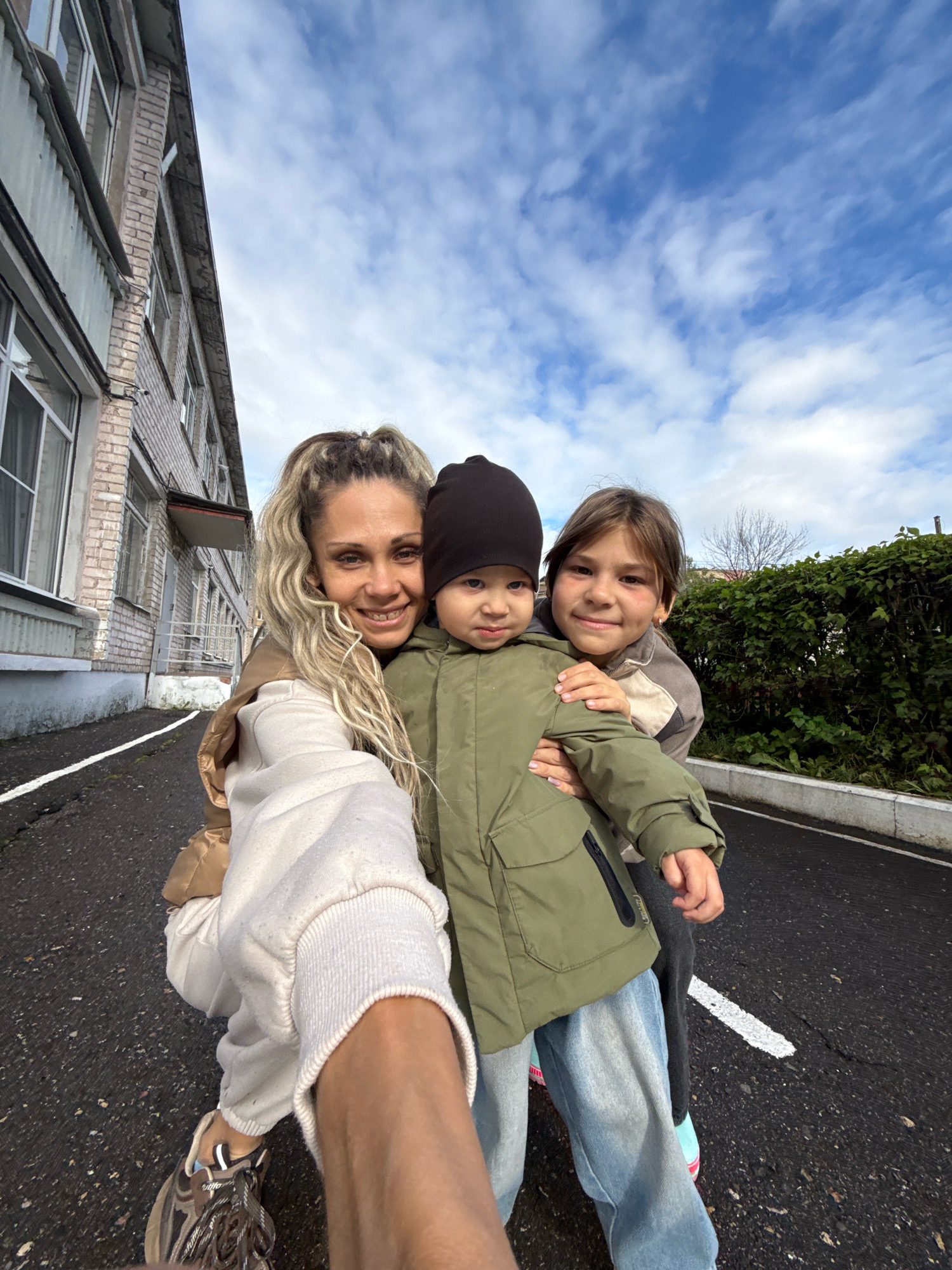 A selfie taken outdoors in Rusanovo, Russia, shows a woman holding two young children. The woman, with blonde, wavy hair, is wearing a beige jacket and a cream-colored sweatshirt. She is smiling and looking at the camera. The child in the center, wearing a black beanie and a green jacket, is being held by the woman and is looking at the camera with a neutral expression. The second child, a girl with brown hair, is standing next to the woman and is also looking at the camera with a slight smile. She is wearing a grey and cream jacket. They are standing on a paved road with a white line marking the edge. A brick building with windows is visible to the left, and a dense green hedge lines the right side of the road. The sky is blue with scattered white clouds.