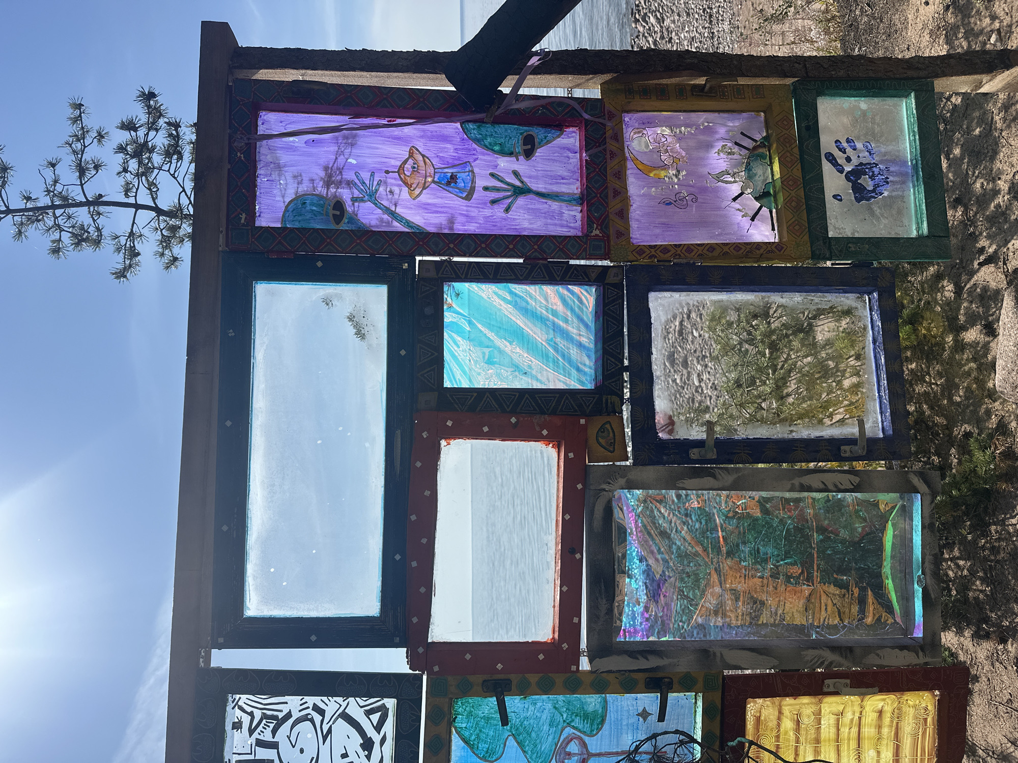 This image depicts an outdoor art installation composed of various repurposed window frames, filled with different materials and artwork. The installation is situated in Luzhki, Russia, near a body of water, likely a lake or sea, with a pebbled shoreline visible. The sky is blue with some scattered clouds and the sun is visible, casting a bright light.
The frames vary in size and color, and are arranged in a somewhat haphazard grid. Some frames contain simple glass, allowing a view of the landscape beyond. Others are filled with colorful, transparent or semi-transparent materials that create abstract patterns and light effects. Several frames feature hand-drawn artwork, including stylized figures, geometric designs, and a depiction of a crescent moon with musical notes. One frame contains a blue handprint. The materials used for the artwork appear to be paint, markers, and possibly cellophane or iridescent plastic.
The installation is constructed from wooden beams and posts, suggesting it is a temporary or rustic structure. A small pine tree grows near the top of the installation, its branches reaching towards the sky. The ground beneath the installation is sandy and covered with small rocks and some sparse vegetation. The overall impression is one of a creative and whimsical roadside art display, likely created by local artists or residents.