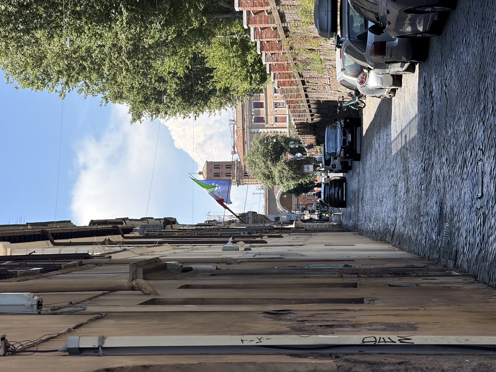 This image depicts a narrow, cobblestone street lined with old buildings on the left and a brick wall with vegetation on the right. A line of parked cars is visible on the right side of the street, extending into the distance. A bicycle is parked near the cars. Overhead, there are power lines and a flag, appearing to be the Italian flag with European Union stars, is flown from a pole attached to one of the buildings. In the background, an arched structure and a tower are visible. The sky is blue with scattered clouds, and trees line the right side of the street. The overall impression is of an urban environment, likely in Italy, with a historic feel.