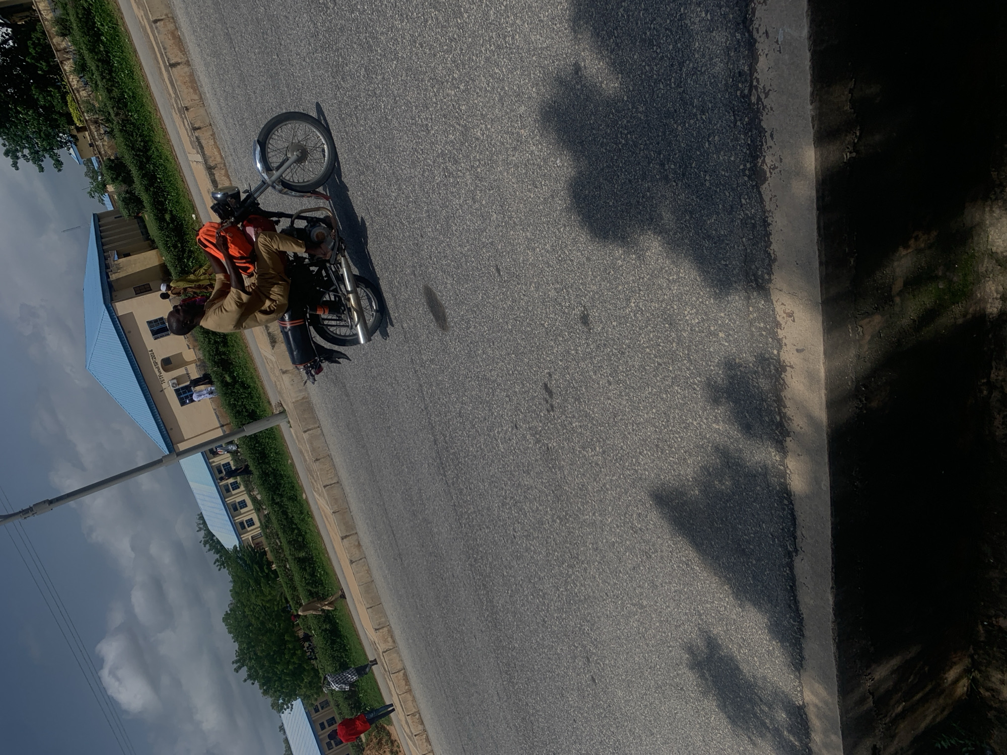 The image shows a person on a motorcycle on a paved road. The motorcycle is red with black accents and has a metal exhaust pipe. The person is wearing a tan uniform and an orange cloth is draped over their shoulders. The scene appears to be outdoors, with buildings and trees visible in the background. The buildings are light brown with blue roofs and have multiple windows. There are also green bushes lining the road. The sky is cloudy, suggesting it might be overcast or that rain is possible. The road is made of asphalt with a concrete curb. Shadows of trees are cast on the road, indicating it is daytime. There are a few other people visible in the background near the buildings. The overall impression is that of an everyday scene on a road in a town or city. There is no discernible text in the image.