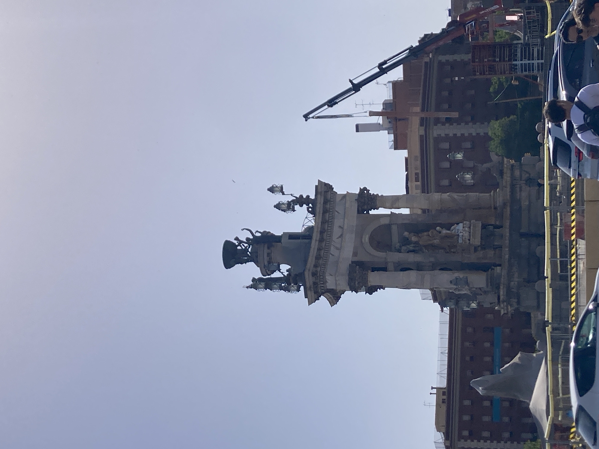 This media file shows an outdoor scene in Barcelona, Spain. The primary subject is a large, ornate fountain with statues, likely a monument or public artwork. It appears to be under some form of construction or renovation, with scaffolding and barriers visible. In the background, a multi-story building with a red brick facade and arched windows is present. A construction crane is also visible extending over the building and the fountain. Several people are in the foreground, some appearing to be construction workers or bystanders observing the scene. The lighting suggests it is daytime, with a clear, bright sky. The overall impression is a busy urban environment with ongoing construction work impacting a historical landmark.