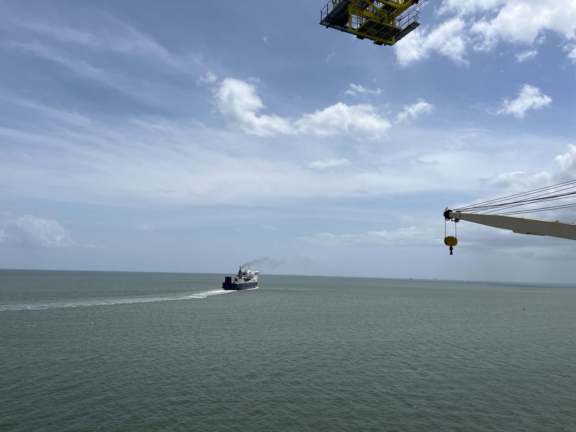 The image shows a large body of water under a partly cloudy sky. A cargo ship is moving across the water, leaving a wake behind it. In the foreground, parts of a crane and its suspension cables are visible. In the top part of the image, there is a structure of another crane.