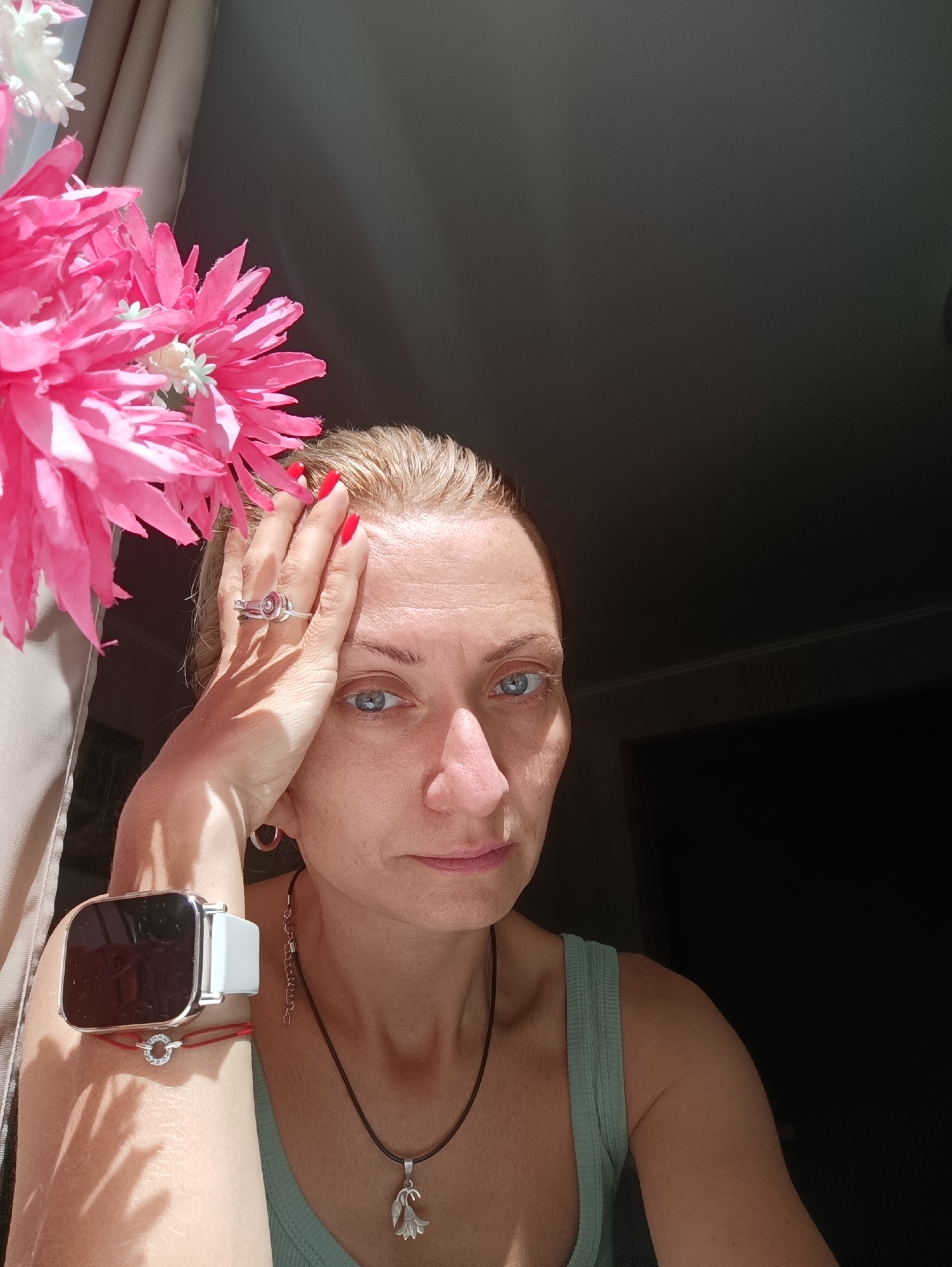 The image shows a close-up of a woman with blonde hair and blue eyes. She is wearing a mint green tank top, a silver ring with a red stone, a smart watch with a white band, and a necklace with a silver pendant. Her right hand is resting on her forehead. In the background, there is a curtain and a dark wall. Artificial pink and white flowers are in the upper left corner of the image. The lighting suggests it is daytime, with sunlight streaming in from the left. The scene is indoors. The location is not specified in the image.