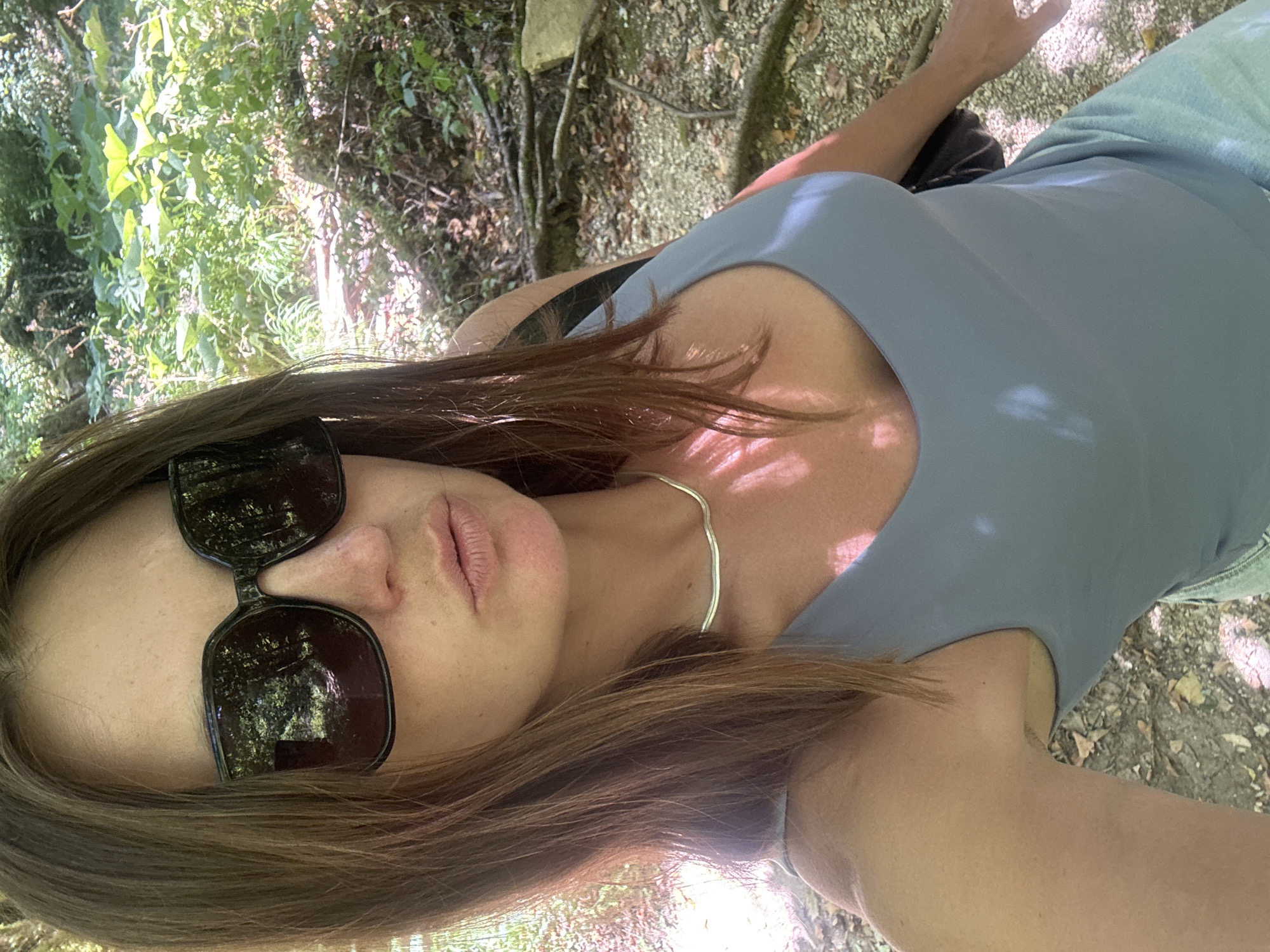 A woman with long brown hair and large dark sunglasses takes a selfie outdoors. She is wearing a light blue tank top and a silver necklace. The background shows a forest with green foliage and dappled sunlight. The ground is covered with dirt, leaves, and exposed tree roots. The location is Coteaux du Lizon, France.
