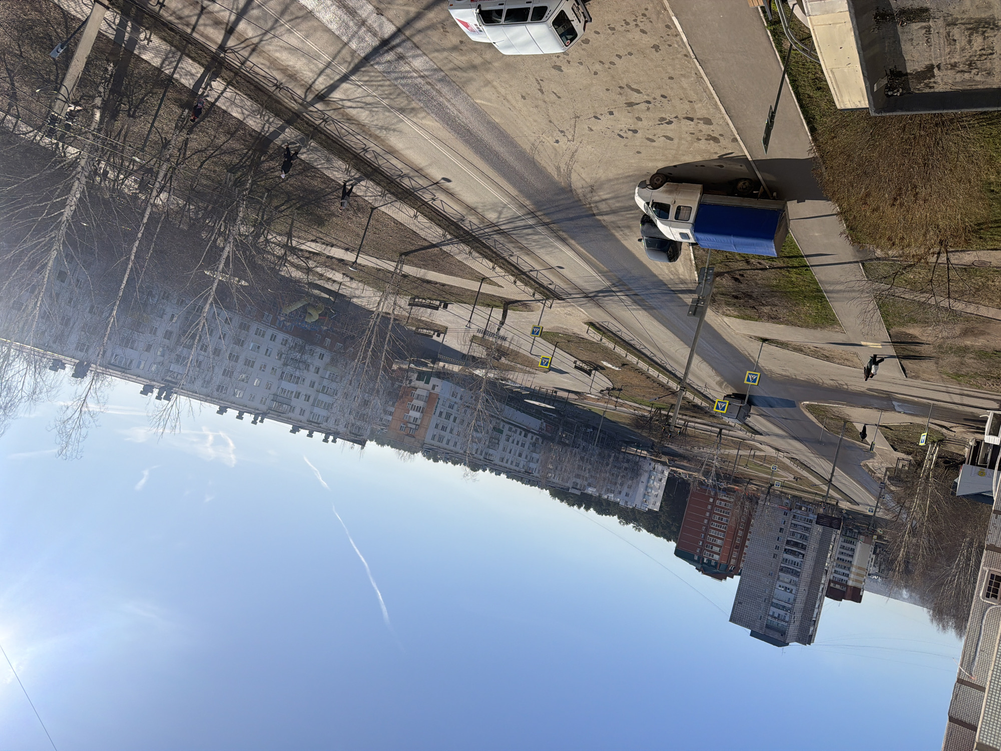 The image presents an elevated view of a street in Chaikovsky, Russia, likely taken from a high-rise building. A clear blue sky dominates the upper portion of the frame. The cityscape includes residential buildings of various heights and architectural styles, primarily in muted colors. Leafless trees are interspersed between the buildings and along the roadside, indicating it is likely early spring.
The street below features pedestrian walkways on either side. Two light-colored vans are parked on the right side of the street, one with a blue tarp covering a portion of its bed. Pedestrian crossing signs are visible at intervals along the road, suggesting a designated crossing area. People are seen walking along the sidewalks and crossing the street. Shadows of trees and overhead wires are cast onto the road surface.