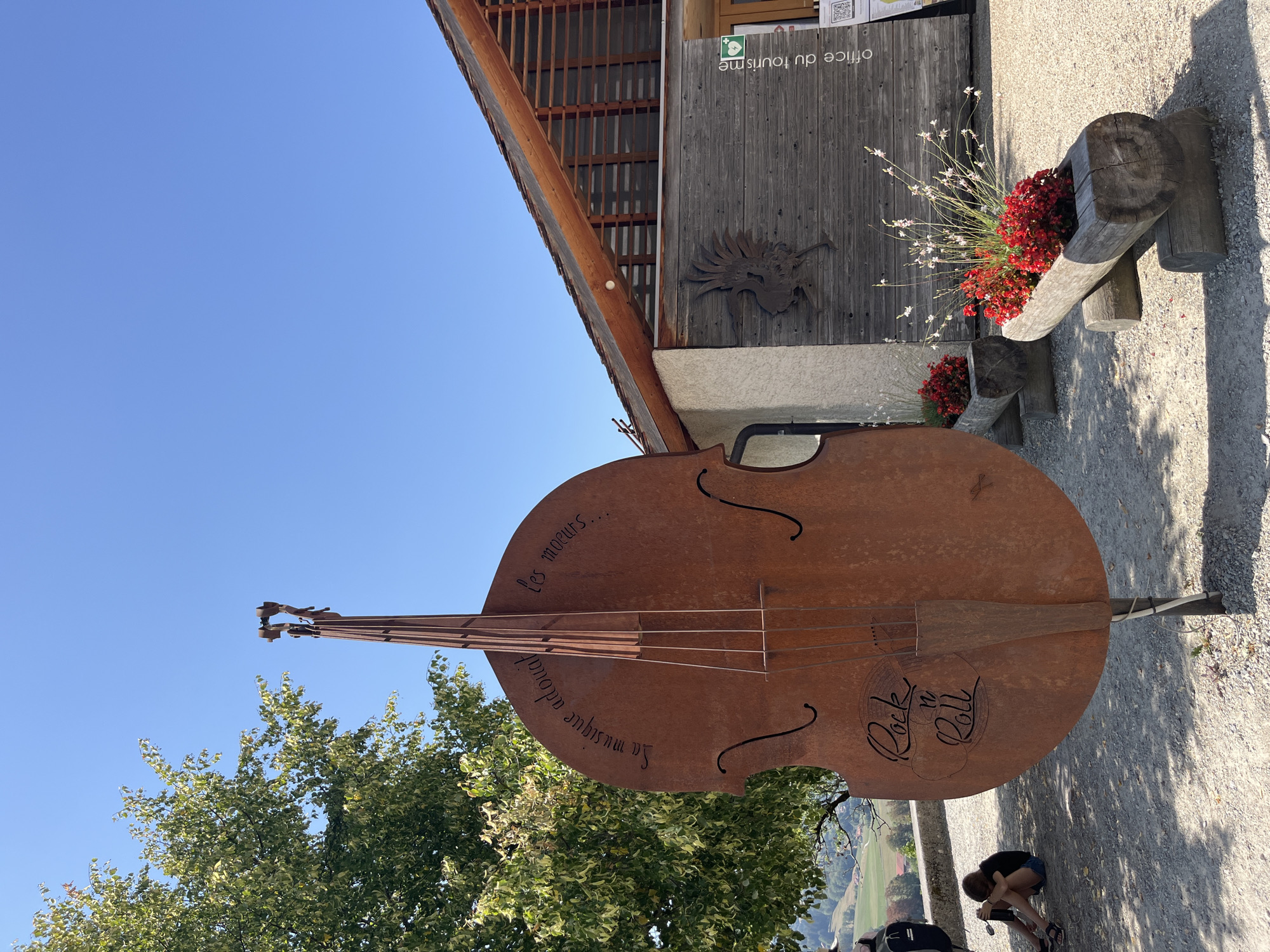 In Epagny, Switzerland, an outdoor scene features a large, rusted metal sculpture of a double bass positioned in the foreground. The sculpture has text inscribed on it, reading "La musique adoucit les moeurs..." and "Rock n Roll." To the left of the sculpture, a young person with dark hair is sitting on the ground, facing away from the camera, looking down at something in their lap. They are wearing a black t-shirt and denim shorts. Behind them, a tree with green and white leaves is visible. To the right of the sculpture, a rustic wooden building, identified as an "Office du tourisme" (tourism office) by text on its facade, is present. The building has a wooden exterior and a sloped roof with exposed beams. A metal bird sculpture is mounted on the wall of the building. In front of the building, two wooden log planters are filled with red flowers. The ground is covered in gravel, and the sky is clear and blue.