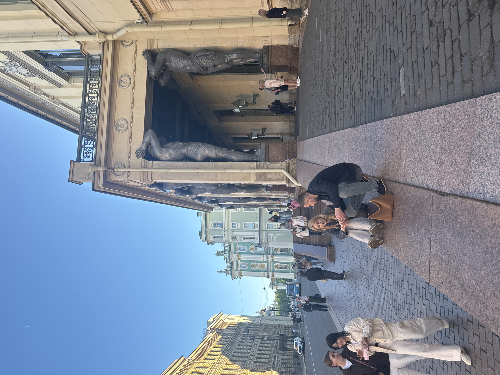 The image captures a street scene in front of the Winter Palace (Hermitage Museum) in St. Petersburg, Russia, under a clear blue sky with a crescent moon visible. Several people are present on the sidewalk and steps leading up to the building's portico, which is supported by large caryatid statues.
In the foreground, two women are walking on the cobblestone sidewalk. One woman, wearing a white jacket and pants, is holding a pink box. The other woman, in a dark jacket and white pants, is looking at her. They appear to be in conversation.
Further back, a young couple is sitting on the stone steps. The woman has blonde hair and is wearing a grey sweatshirt and light-colored pants. The man next to her is wearing a dark jacket and jeans. They are facing each other and interacting.
On the portico level, several individuals are standing and walking. Two people, a man and a woman in a short dress, are near one of the caryatid statues, possibly taking a photo or admiring the architecture. A blonde woman in a black jacket and dark pants is standing to the right, facing away from the camera.
In the background, the distinct green and white facade of another building, likely part of the Hermitage complex, is visible. Cars and a blue bus are on the street, and other pedestrians are scattered along the sidewalk. The overall atmosphere suggests a pleasant day in a historic urban setting.