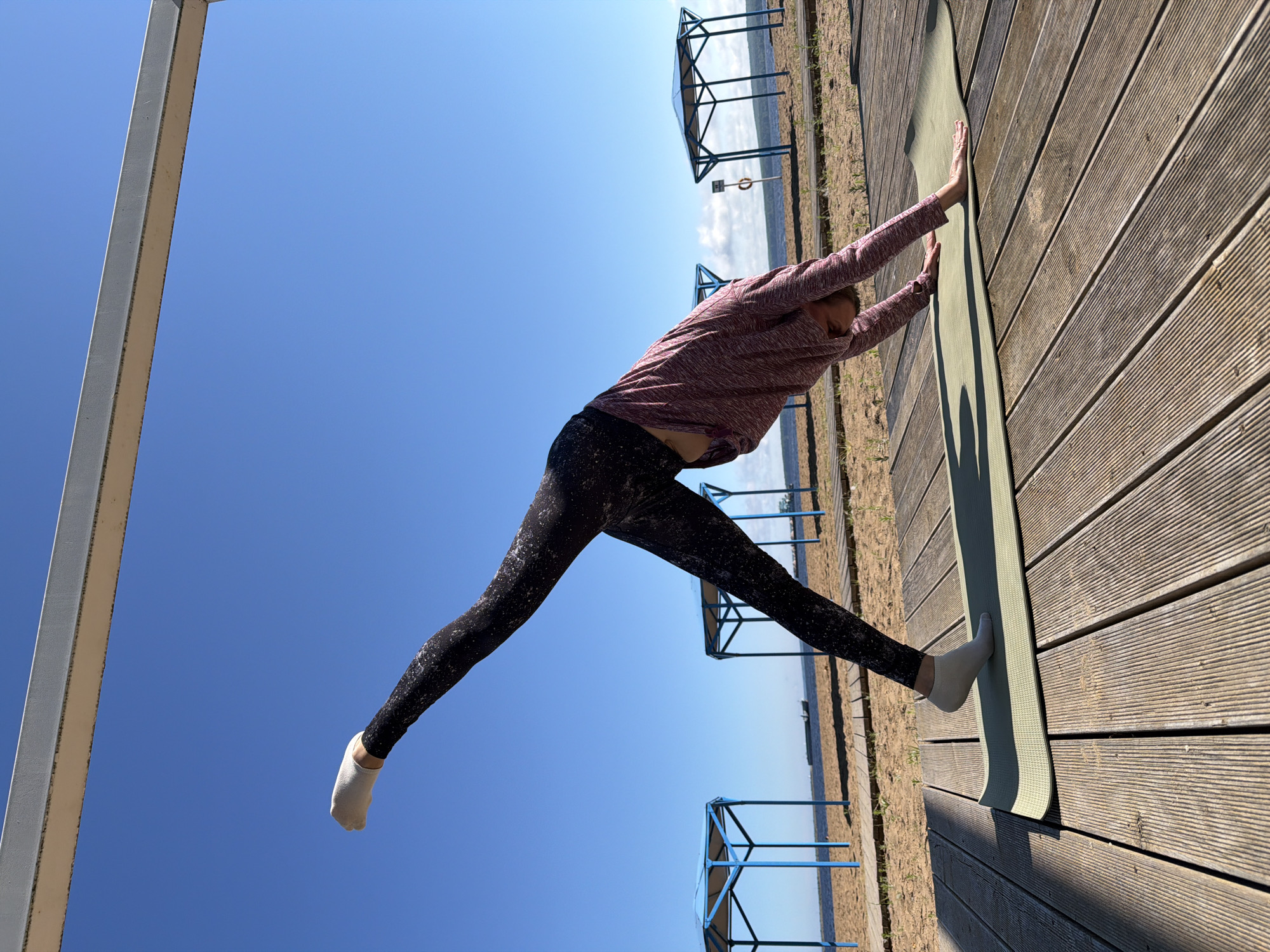 An adult female is performing a yoga pose on a wooden deck at a beach in Chaikovsky, Russia. She is positioned on a green yoga mat, with her body in a downward-facing dog pose with her right leg extended upwards. She is wearing a pink long-sleeved shirt and patterned black leggings. The background shows a sandy beach, the Kama River, two blue gazebos, and a clear blue sky. A light-colored structure is visible overhead.