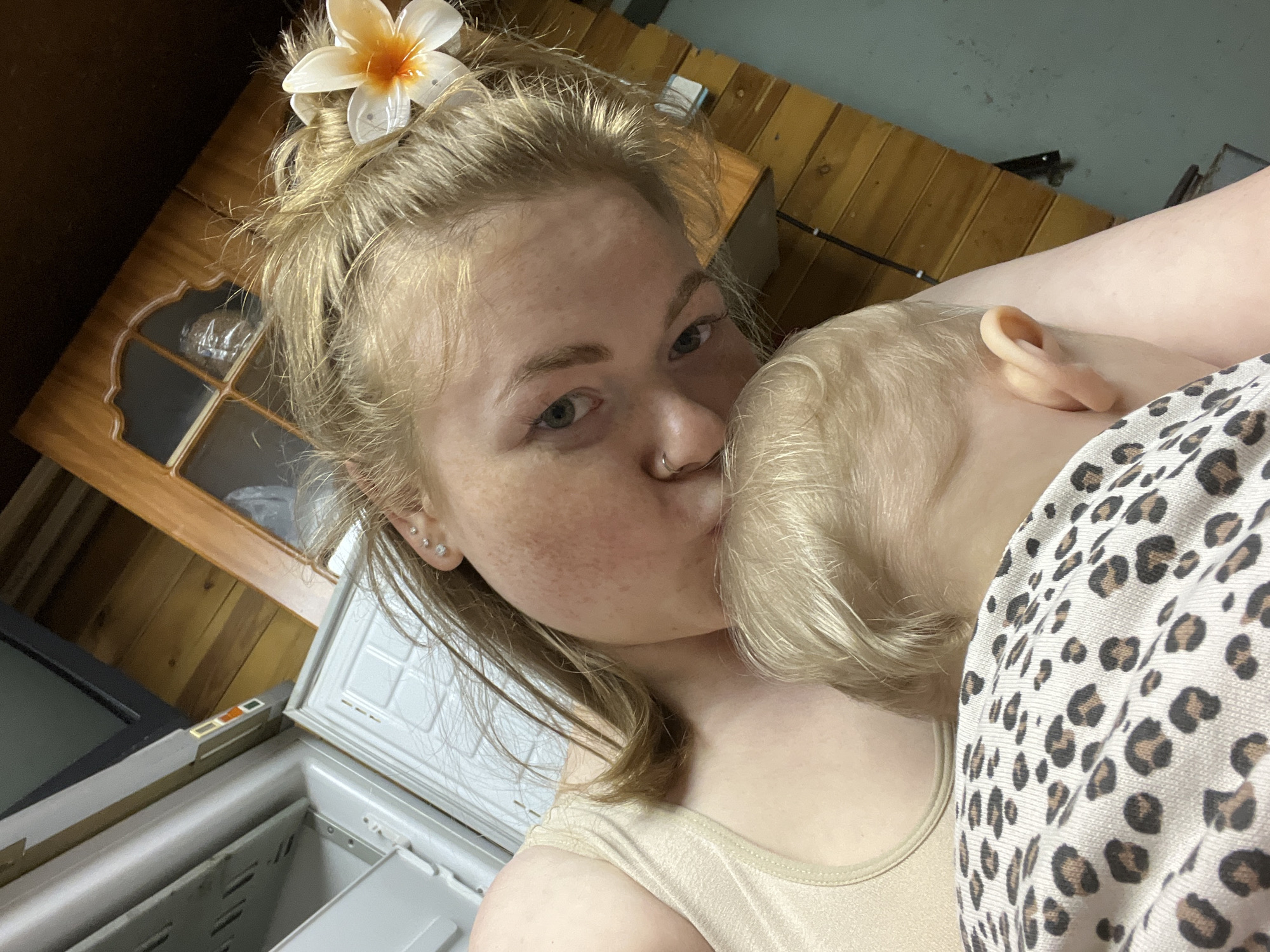 A woman with blonde hair and a flower in her hair is holding and kissing a baby with blonde hair. The woman has a nose ring and freckles. The baby is wearing a leopard-print garment. They appear to be indoors, near a refrigerator, a wooden cabinet, and a wooden wall. The location is likely in Chaikovsky, Russia, based on the request.