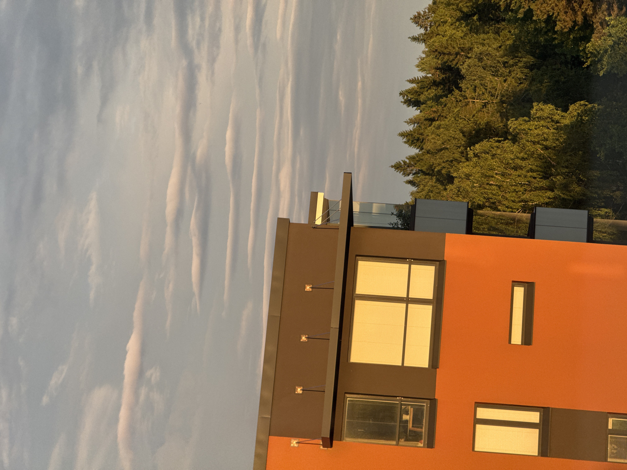 A medium shot captures the side of an orange and grey modern building, with a lush green forest bordering it in the background. The building features several windows, some reflecting the golden light of the setting sun. The sky above is a soft blue, filled with streaks of pale pink and grey clouds. The overall scene evokes a sense of peaceful coexistence between urban architecture and nature.