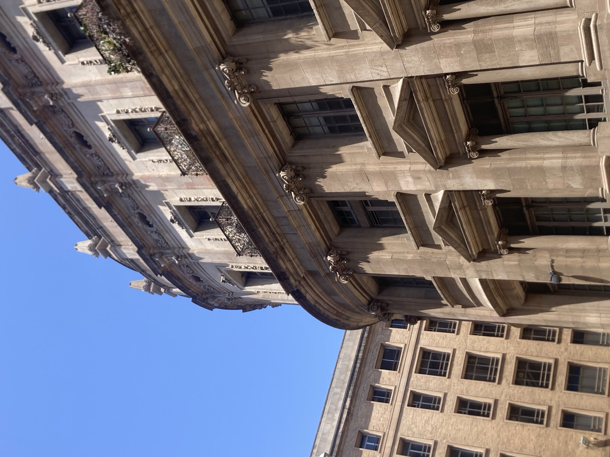 This media file shows an upward-looking view of architectural details of buildings against a clear blue sky. The main subject is the ornate stone facade of a building on the left, with multiple windows and decorative elements. Balconies with intricate wrought iron railings are visible, and some greenery appears to be growing on one of the balconies. The stone work features carved details, pilasters, and projecting cornices.
In the background and to the right, another building with a more modern facade is visible, characterized by rows of regularly spaced windows. This building appears to be made of brick or a similar material.
The lighting suggests it is daytime, with strong shadows indicating sunlight. The clear blue sky implies fair weather. The architectural style of the building on the left, with its classical elements and decorative flourishes, suggests it could be an older, historic building, possibly in a European city. Given the location context of Barcelona, Spain, it's plausible that these buildings are situated in a historic district of the city. No people are visible in the frame. No text is discernible in the image.