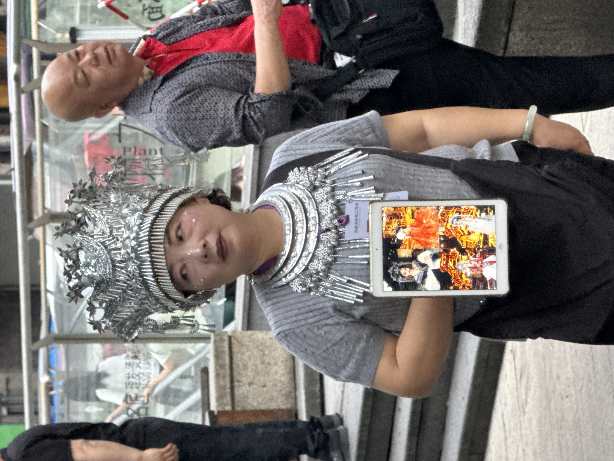 A woman in traditional silver headdress and necklace holds a tablet displaying images of people in similar attire. Behind her, a bald man in a grey shirt and red undershirt lies down. The location appears to be a public area in Yuzhong District, China, with stairs and glass partitions visible. A person wearing black pants and shoes is visible in the foreground.