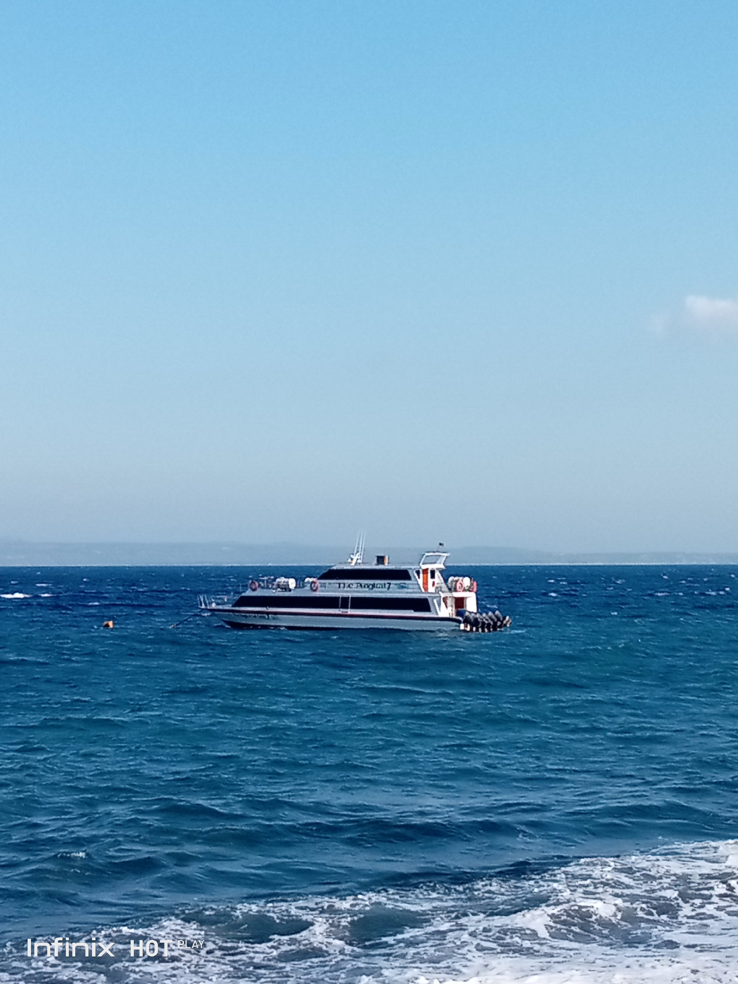 A passenger boat, identified by the text "The Angelic 7" on its side, is anchored in the blue ocean. The boat is white with a dark stripe along its hull and features multiple outboard motors at its stern. A small orange buoy floats in the water to the left of the boat. The horizon displays a faint outline of land under a clear blue sky. The foreground shows choppy water with white foam near a sandy shore. The text "Infinix HOT PLAY" is visible in the bottom left corner of the image, indicating the device used for capture.