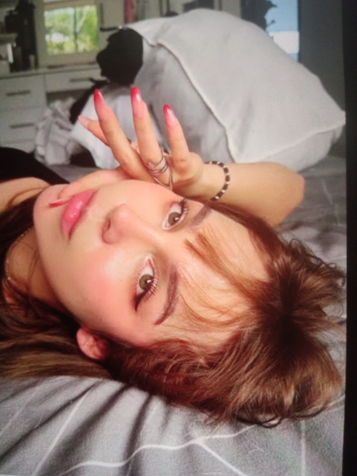 A person with reddish-brown hair and light skin lies on a gray and white striped bed. Their face is close to the camera, tilted upwards, with their left cheek resting on their arm. They are wearing dark clothing on their left shoulder. Their right hand is raised, with the index and middle fingers extended in a peace sign, adorned with long, pointed, gradient red and pink nails. The person wears two silver rings on their left ring finger and a bracelet with dark and white beads on their right wrist. Their makeup includes winged eyeliner and pink blush. The background shows a white pillow, furniture with drawers, and a window with greenery visible outside, suggesting an indoor setting. The image's quality is slightly grainy, and it is unclear if this scene is located in Jalingo, Nigeria, as no specific indicators of this location are present.