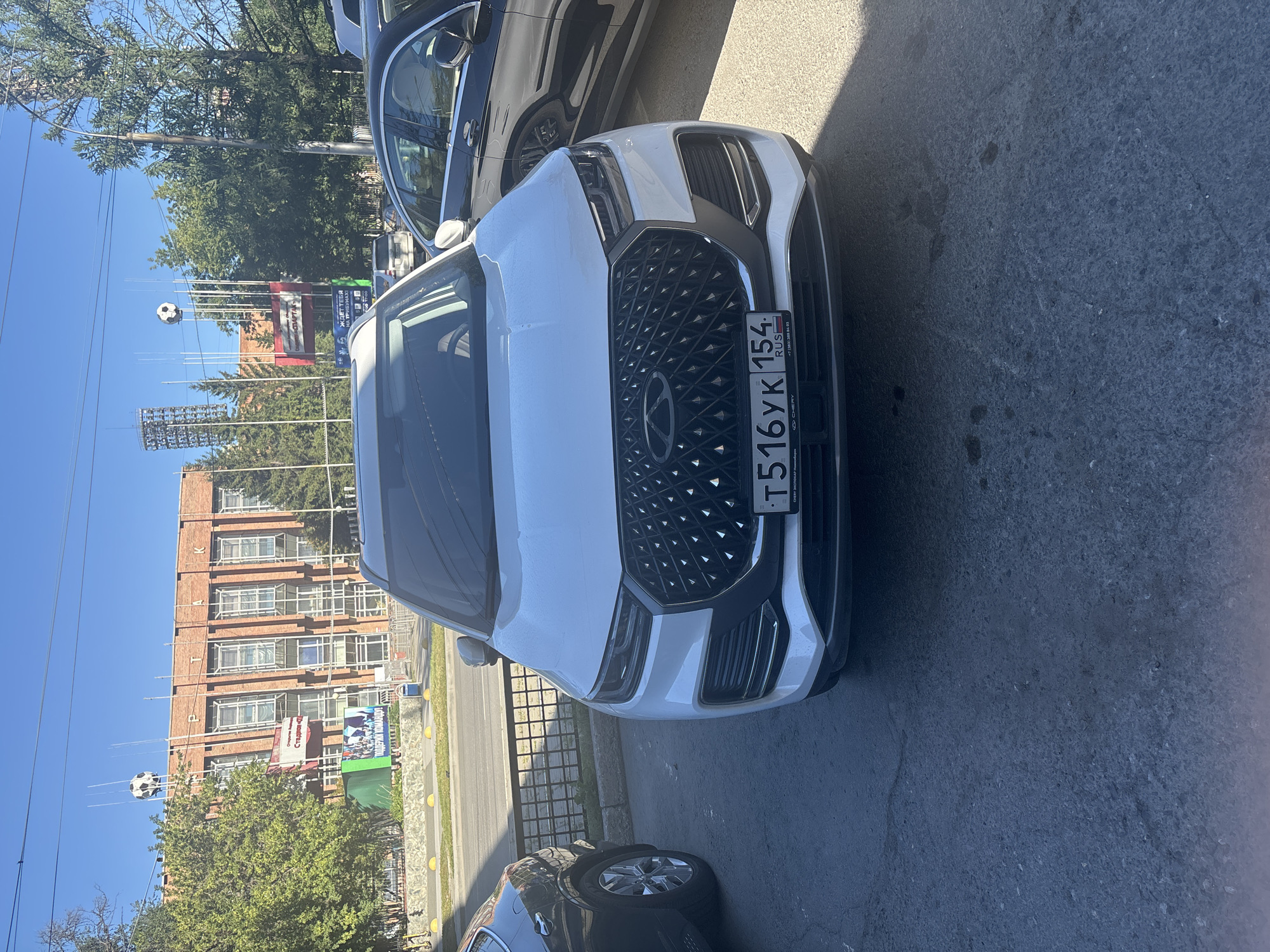 The image shows a white Chery Tiggo 7 Pro car parked on an asphalt surface in front of a brick building and a street in Novosibirsk, Russia. The car's license plate reads "T516YK 154 RUS". In the background, a brick building with large windows is visible, with the word "СПАРТАК" (Spartak) written on it. Several tall poles with what appear to be soccer balls on top are erected near the building. A black Mercedes-Benz sedan is parked to the right of the Chery. The environment suggests an urban setting, possibly near a sports facility. The sky is clear and blue, indicating a sunny day.