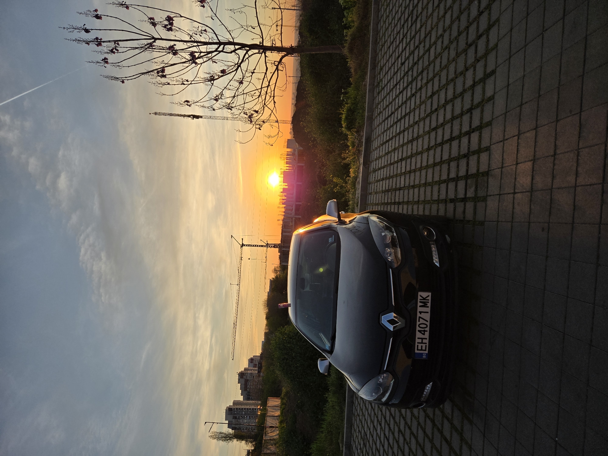 The image, oriented vertically, captures a vibrant sunset over an urban landscape with a car in the foreground.
In the immediate foreground, a dark-colored Renault car is parked on a patterned paved surface, angled slightly towards the viewer. The front of the car, including its headlights, grille, and the Renault logo, is clearly visible. The license plate reads "EH 4071 MK" with "BG" indicating Bulgaria. The car's surface reflects some of the orange glow from the sunset.
Behind the car and extending across the middle and upper parts of the frame, a dramatic sunset dominates the sky. The sun itself is visible as a bright, intense disk on the horizon, casting a strong orange and yellow hue across the sky with wispy clouds. Several tall construction cranes and numerous buildings are silhouetted against the bright sky, indicating an urban or developing area. To the left side of the frame, a bare-branched tree with some small, dark reddish flowers is silhouetted against the sky. A faint white contrail from an aircraft stretches across the upper left portion of the sky.
The scene suggests late afternoon or early evening. The overall atmosphere is scenic, contrasting the natural beauty of the sunset with the industrial elements of cranes and buildings, and the parked vehicle.