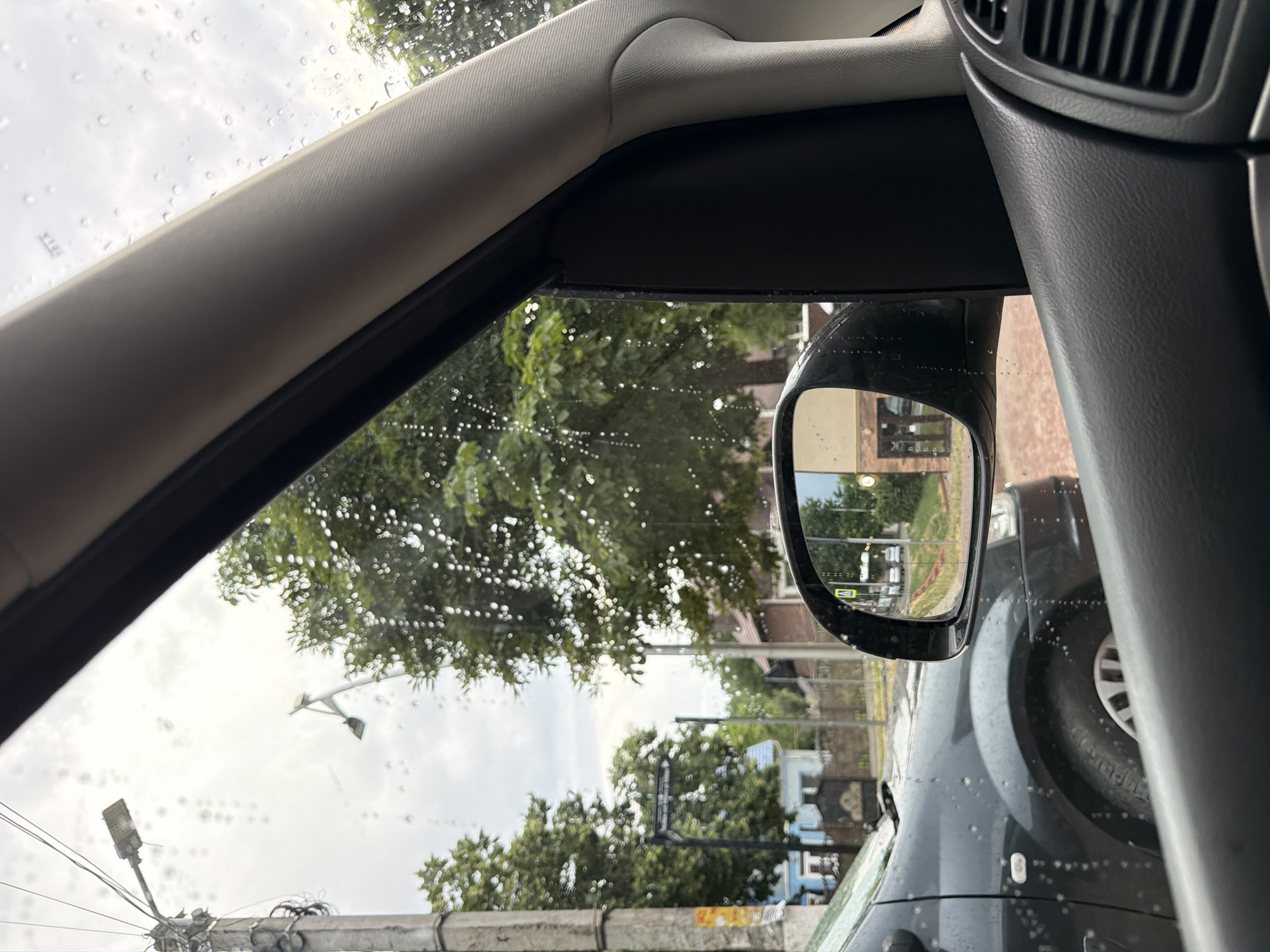 The image, captured from inside a vehicle, shows a view through the passenger-side window. The window is wet with raindrops, creating a blurred effect on the external scene. In the foreground, the interior of the car is visible, including a dark gray door panel with a textured surface and a part of the air vent. To the left, a portion of the car's exterior, a dark gray vehicle, is visible.
The exterior scene includes a concrete utility pole on the left, with electrical wires and a street light attached. Further back, trees with green foliage are prominent. A street sign is visible hanging from a pole. In the background, there are several houses, one with a light blue facade and a dark gate, and another with a red brick facade. A side mirror of the car reflects a street scene with buildings and parked vehicles, suggesting a street in Strășeni, Moldova. The sky appears overcast, with clouds and diffused light. No people are visible in the image. The overall impression is a view of a residential street in Strășeni during a rainy day.