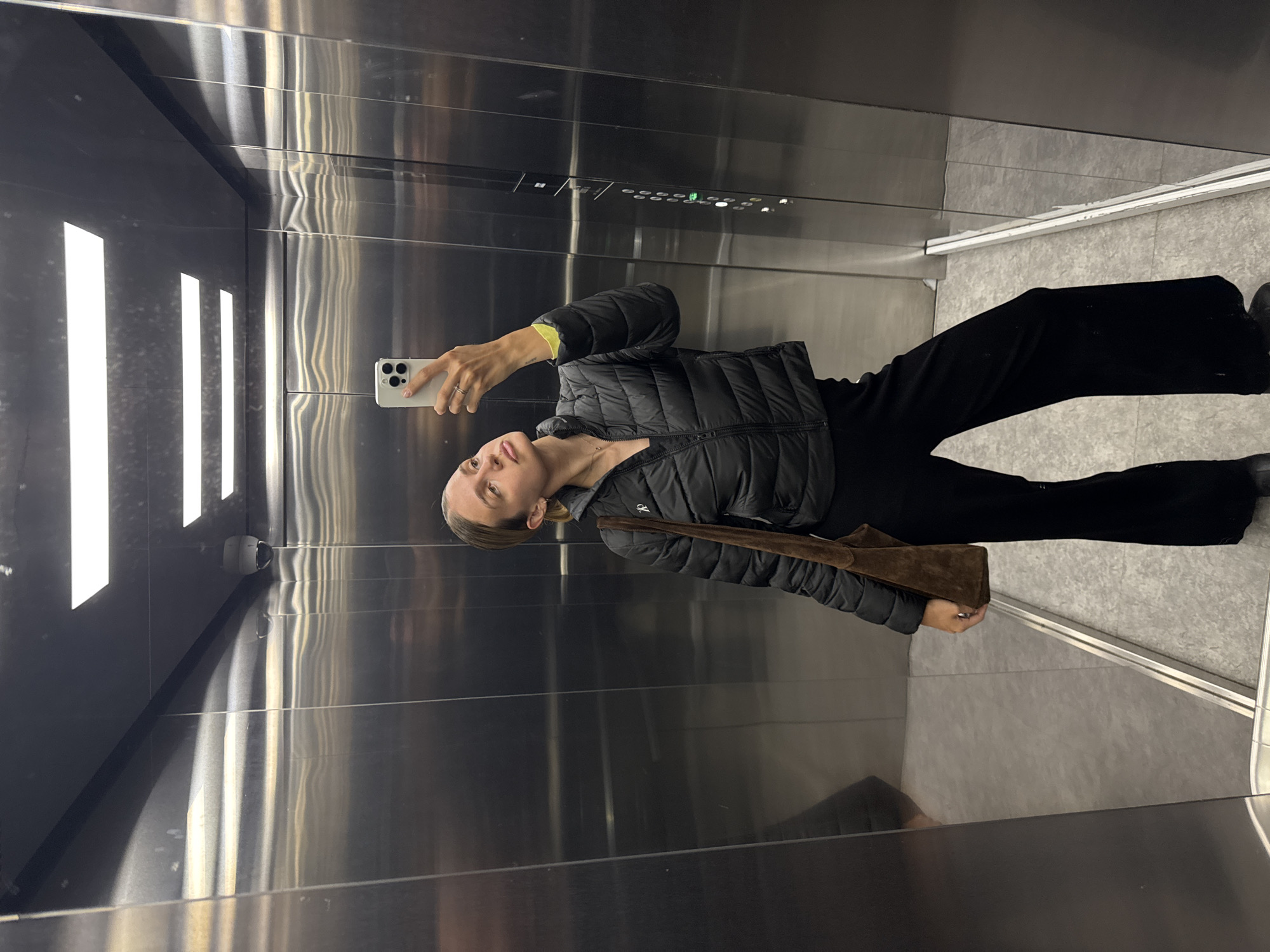 A woman is taking a selfie in an elevator. She is wearing a black puffer jacket, black pants, and holding a brown purse. The elevator has a stainless steel interior and the floor is grey. There are long, vertical lights on one wall. A security camera is visible in the top corner of the elevator.