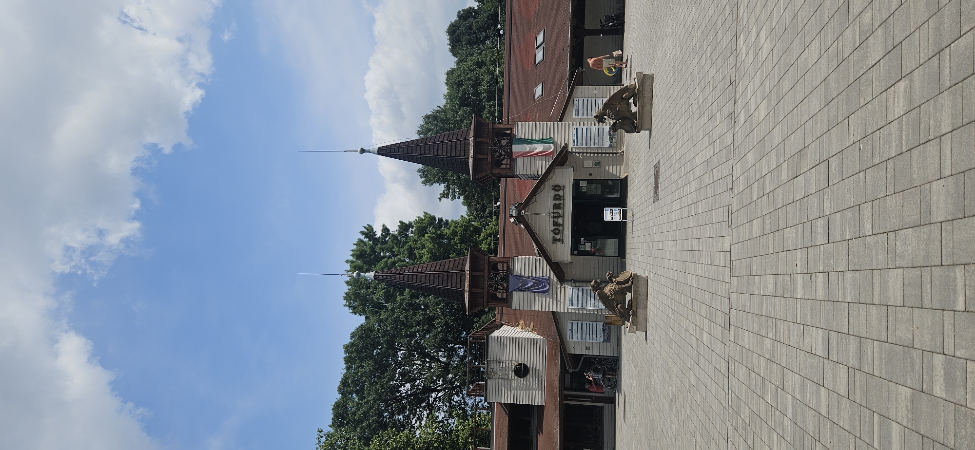 The image shows the entrance to the Hévíz Thermal Bath in Hévíz, Hungary, on a sunny day with scattered clouds. The building features a traditional architectural style with two prominent pointed towers topped with lightning rods. Flags hang from the building, including the Hungarian flag. In front of the entrance are two stone sculptures of winged lions. The paved area in front of the building is clean and well-maintained. A few people are visible in the scene, including a man in shorts carrying a yellow ring near the right side of the building and a couple with bicycles near the left side. The environment is outdoor, with trees visible behind the building, suggesting a park-like setting.