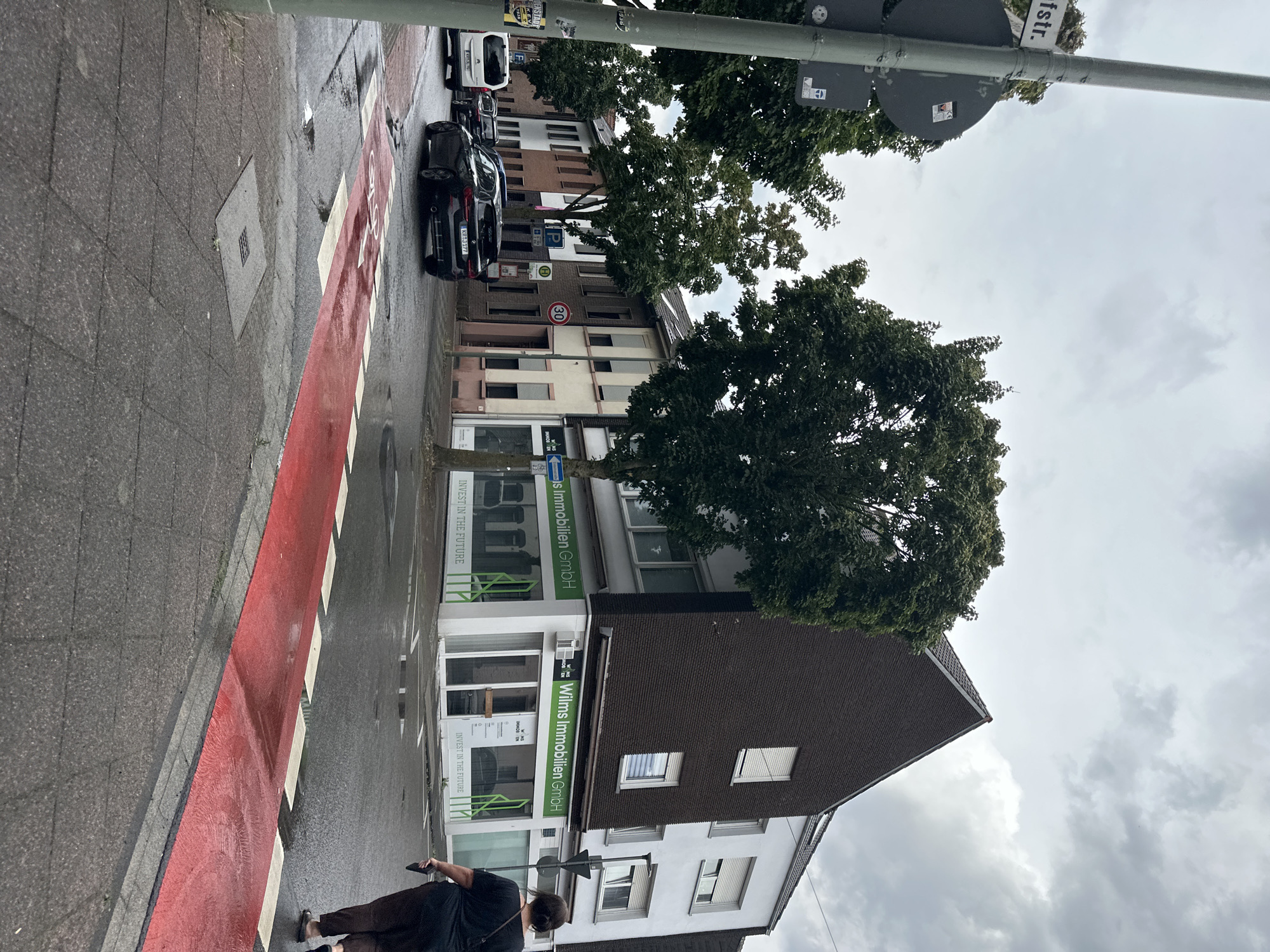 The image depicts a street scene in Krefeld, Germany, on a cloudy day. A red-painted bicycle lane with white dashed lines is visible in the foreground, leading across a wet street. A black BMW sedan with license plate KR J 177 is stopped on the street. To the right of the bicycle lane, a woman with dark hair, wearing a dark t-shirt and patterned pants, walks away from the camera.
In the background, a building with a dark brick facade and a white upper section houses "Wilms Immobilien GmbH" on its ground floor, with green signage and text that reads "INVEST IN THE FUTURE." Adjacent to this building is a white building. Across the street, a row of smaller buildings lines the street, with a "30" speed limit sign visible on one of them. A white van is parked on the left side of the street. Trees with green foliage are interspersed throughout the scene. A street sign on the left reads "Bistr."