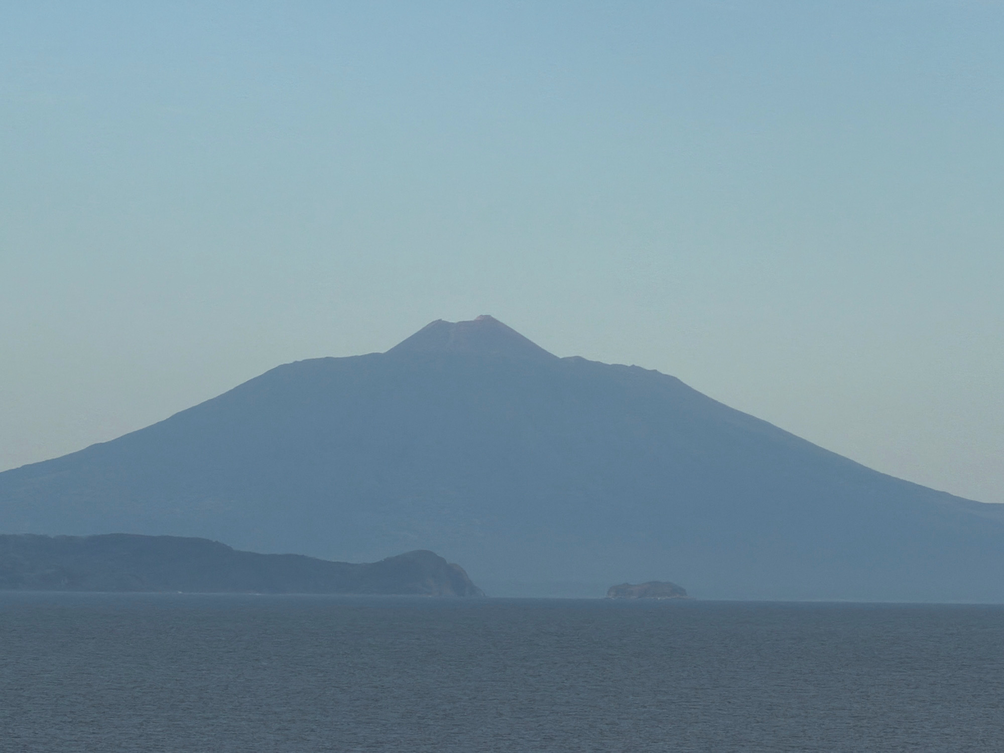 A wide shot captures a vast, hazy blue mountain range rising from a calm, dark blue sea under a pale blue sky. The largest mountain, a dormant volcano with a visible crater at its peak, dominates the center of the frame. In the foreground, the ocean stretches across the bottom half of the image, with a small, rocky island visible in the middle ground to the right. The distant shoreline features a series of smaller, rolling hills. The scene appears serene and unpopulated, with no signs of human activity.